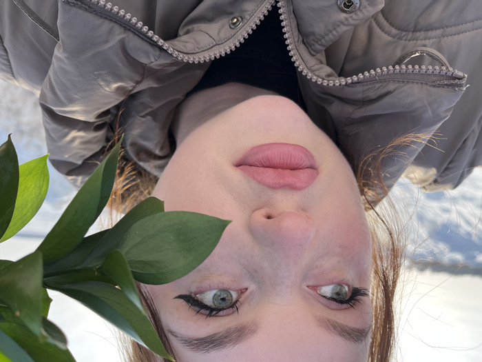 A close-up photograph depicts a young adult female subject outdoors in Pavlovka, Russia, against a snowy landscape under bright sunlight. The subject has reddish-brown hair, fair skin with visible freckles and flushed cheeks, light green eyes, and full lips with light pink lipstick. Her left eye (viewer's right) is fully visible, featuring black winged eyeliner and prominent eyelashes, while her right eye (viewer's left) is partially obscured by a cluster of vibrant green leaves from an unidentified plant branch held in the foreground. She is observed looking upwards and to her left (viewer's right). The subject wears a light brown or beige puffer jacket over a black garment, with a zipper visible on the jacket's front. The background consists of an expanse of snow-covered ground extending to a distant horizon under a clear, bright sky, indicative of a sunny winter day.