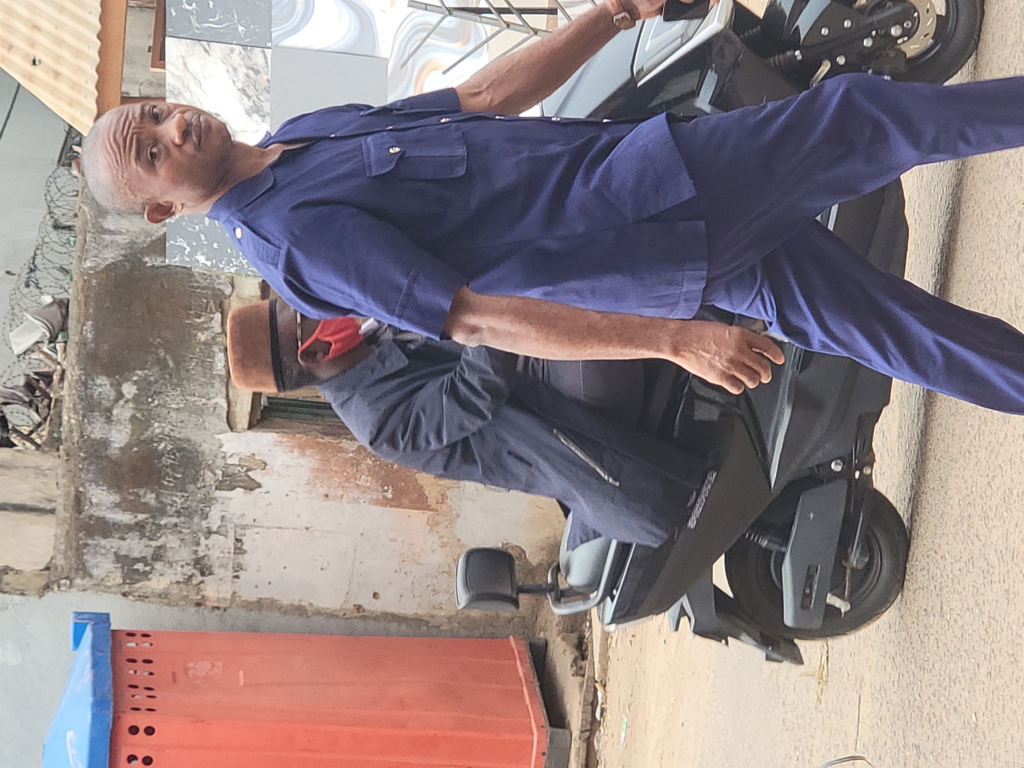 A man in a blue uniform is seated on a black motorized scooter in an urban environment. Another man wearing a brown hat and a red face mask stands next to the scooter. The setting appears to be Maiburiji, Nigeria. The background shows buildings with weathered facades and a concrete curb. There is a red and blue object in the lower left corner. The scene suggests transportation or mobility within the urban area.