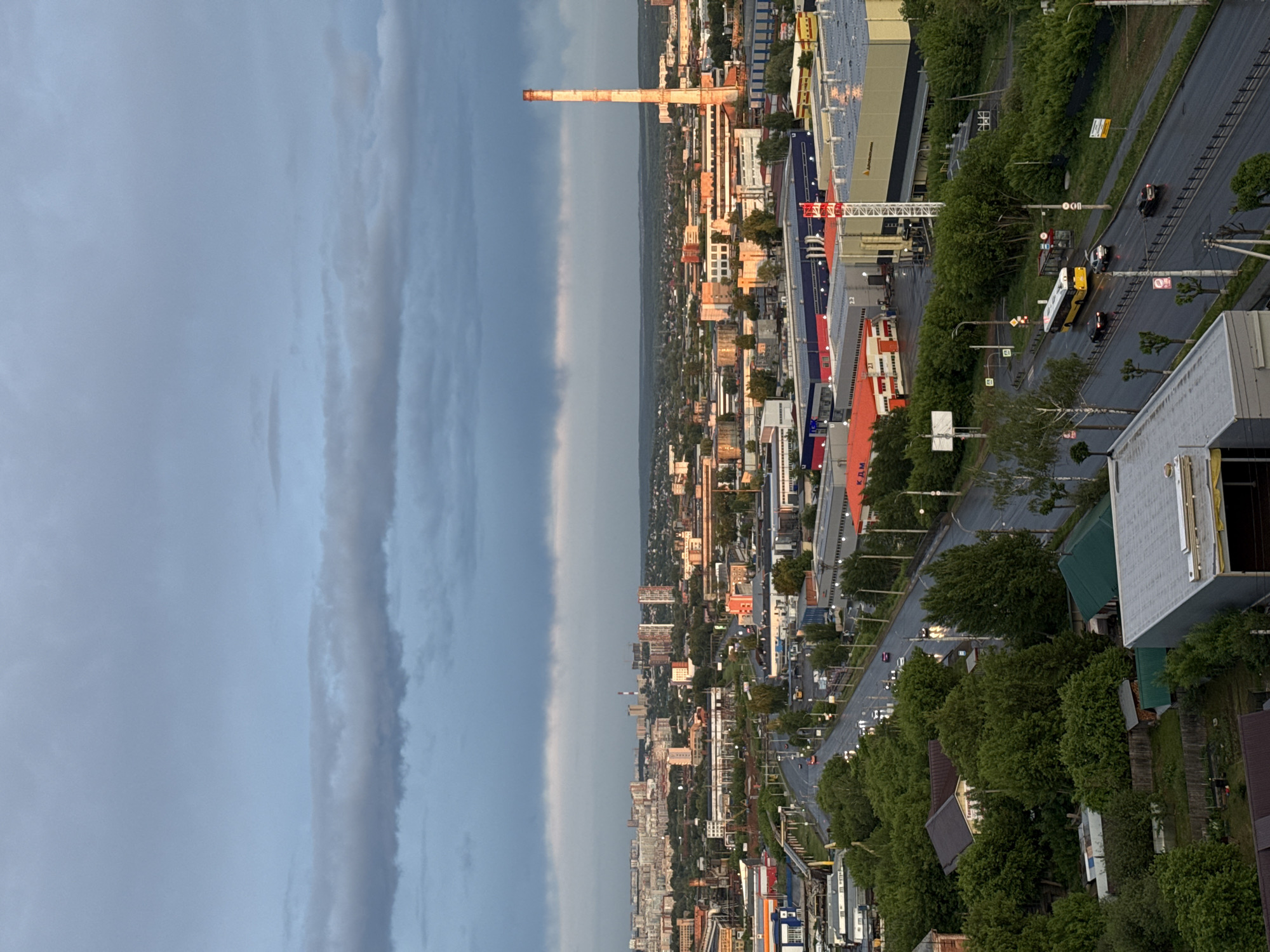 This image shows a panoramic view of Izhevsk, Russia, from an elevated perspective. The sky is overcast with striated cloud formations. Below, the cityscape is revealed, featuring a mix of residential and industrial buildings. A prominent industrial chimney stands tall, its brickwork distinct. Roads stretch through the urban landscape, showing moving vehicles, including cars and a yellow bus. The streets are lined with trees. The tops of some buildings are visible near the viewpoint.