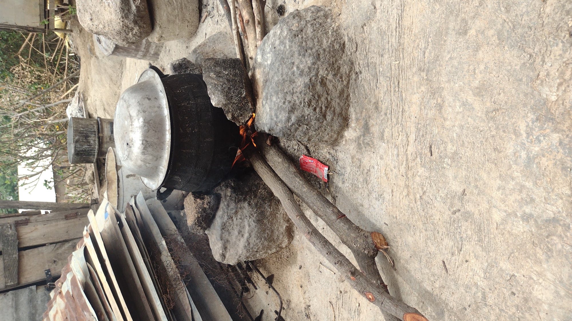 The image depicts an outdoor cooking scene set in what appears to be Jos, Nigeria, during the daytime. A large, dark metal pot with a shiny metallic lid is positioned over an open fire, supported by several rocks that act as a makeshift stove. Wooden branches are used as fuel, with one visibly extending into the active flames.
The ground is concrete or cement, displaying a textured, worn surface. To the left of the cooking area, a stack of corrugated metal sheets and wooden planks are visible, along with another smaller metal container or pot in the background. Sparse, possibly leafless, tree branches and a glimpse of a building structure can be seen in the upper left background, suggesting a semi-urban or backyard environment. The bright, even lighting indicates clear weather.
A small red wrapper or sachet lies on the ground near the fire; it has white text on it, but the details are too blurry to be deciphered. The scene conveys a practical and traditional method of preparing food outdoors.