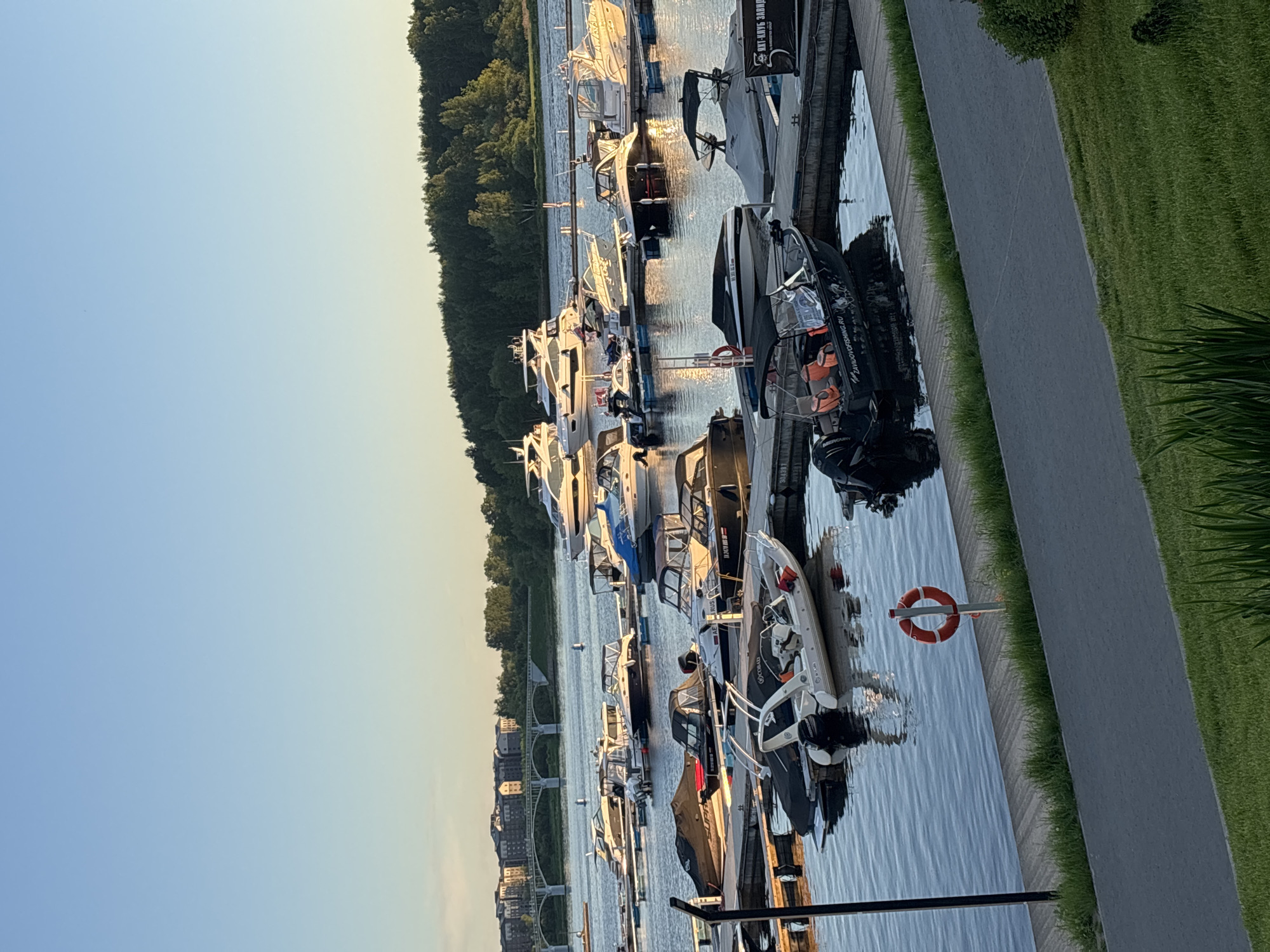 The image depicts a marina in Varaksino, Russia, at dusk. Several boats of varying sizes and designs are docked along a concrete pier adjacent to a paved walkway and grassy area. A forested area lines the opposite side of the waterway, with buildings visible in the far distance. An orange life preserver is mounted on a pole near the pier's edge. The sky is clear and transitioning from blue to a lighter color with the setting sun.