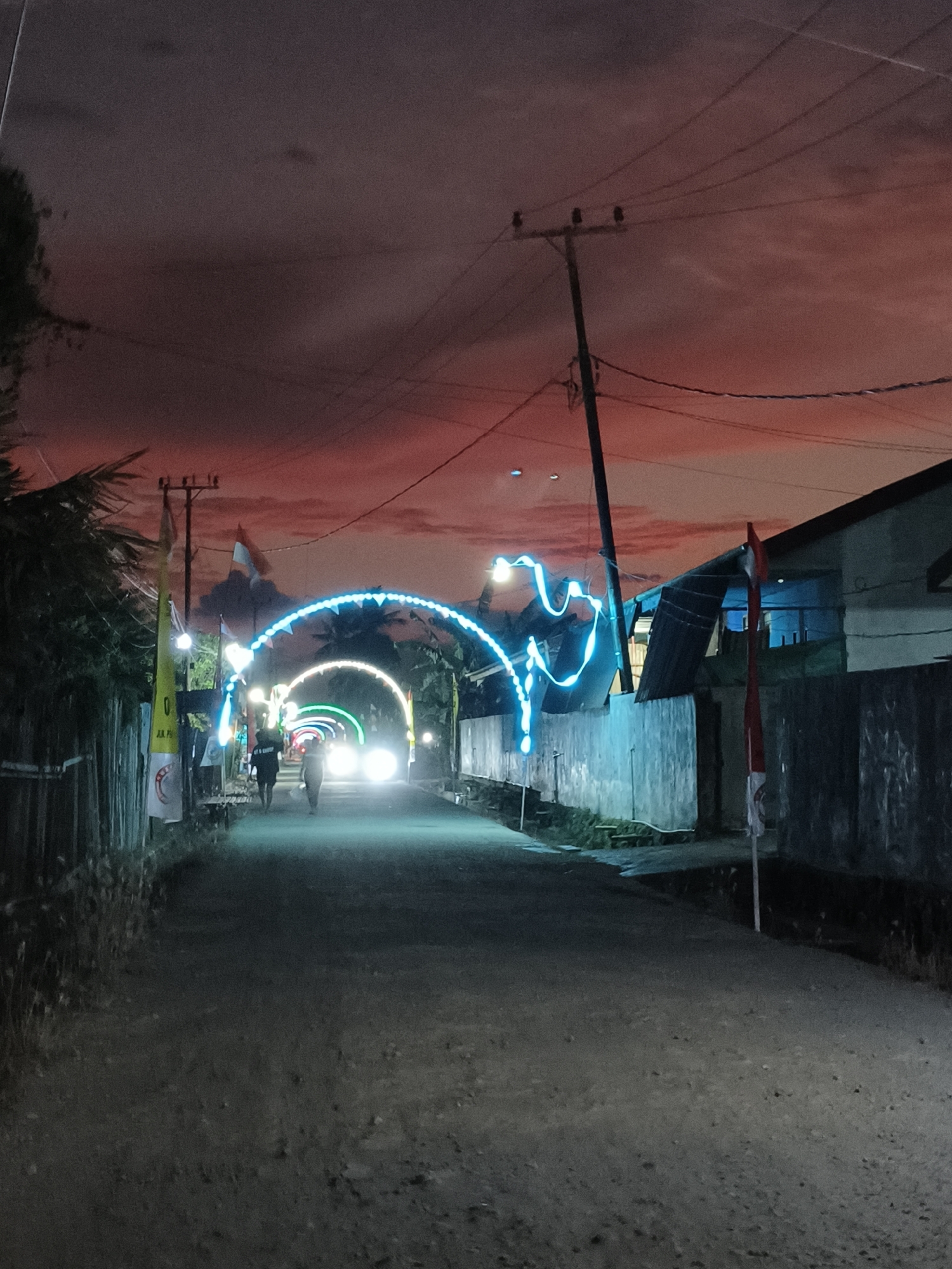 This image captures a street scene at dusk in Mariat Pantai, Indonesia, with a dramatic red sky overhead. The street is lined with a fence on the left and a building on the right. Two figures are walking away from the camera down the road. Archways decorated with blue and green lights span across the street, suggesting a festive or celebratory atmosphere. Several flags are visible, including Indonesian flags. A utility pole with wires runs overhead. The road appears to be unpaved and somewhat rough. The overall lighting suggests it is either early morning or late evening.