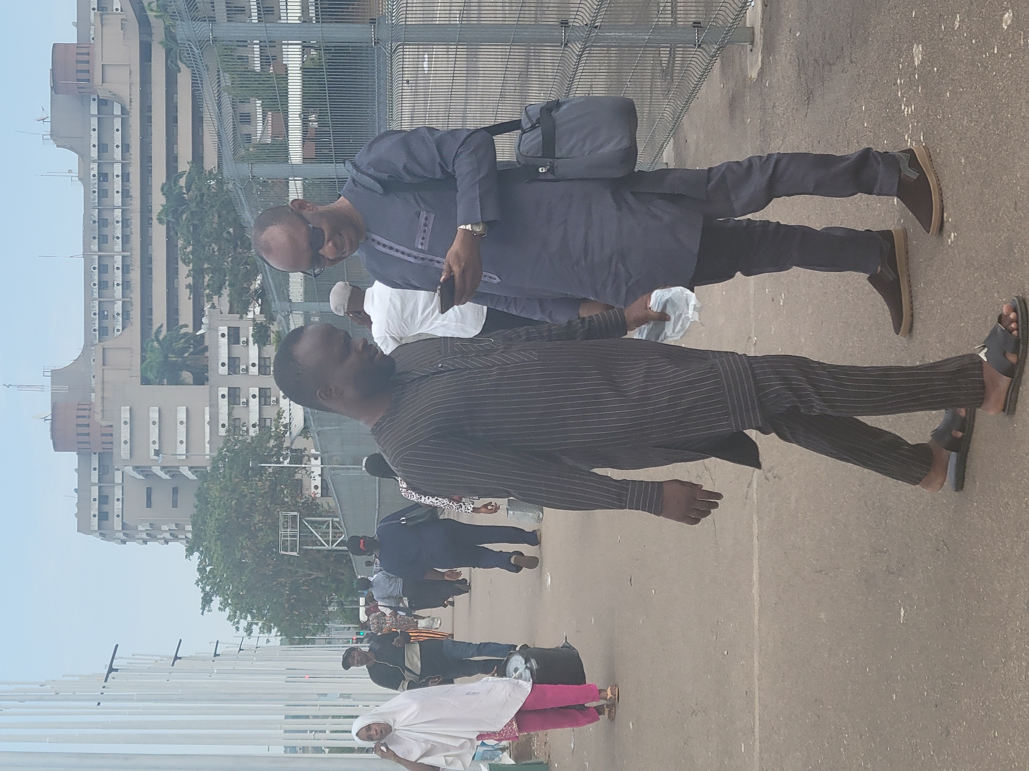 The image shows a public walkway in Abuja, Nigeria. A group of people are walking on the sidewalk. Two men are in the foreground. The man in front is wearing a striped shirt and black slacks with sandals. The second man is wearing sunglasses, a long blue shirt, and black slacks with brown shoes. He is holding a smartphone in his right hand and a bag in his left. A tall, multi-story building can be seen in the background. The building has a light brown exterior with repeating vertical columns. A metal fence is visible on the right side of the walkway. The sky is overcast and a pale blue color.