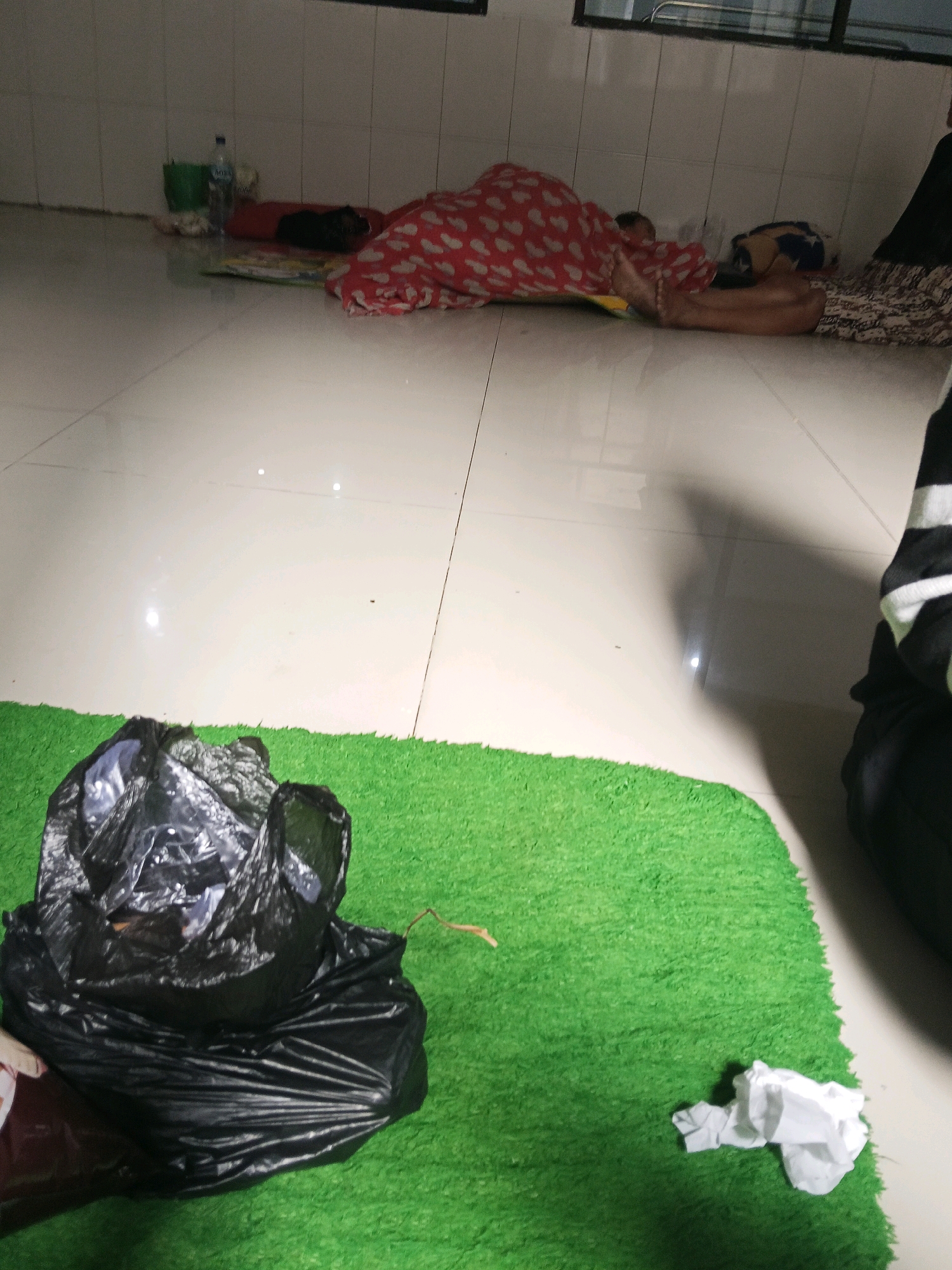 In Kediri City, Indonesia, the image depicts a tiled floor within an indoor space, possibly a waiting area or ward, with tiled walls. In the foreground, a bright green artificial grass mat is visible, on which a large, crumpled black trash bag lies. To the right of the trash bag, a crumpled white tissue is also present on the green mat. Further back, two individuals are resting or sleeping on the floor. One individual is covered with a red blanket patterned with white hearts, lying on their side. Another person is partially visible next to them, also lying down. In the background, near the tiled wall, several items are scattered, including a plastic water bottle, a green bag, and other indistinct objects. The lighting suggests it is either evening or the room is dimly lit.