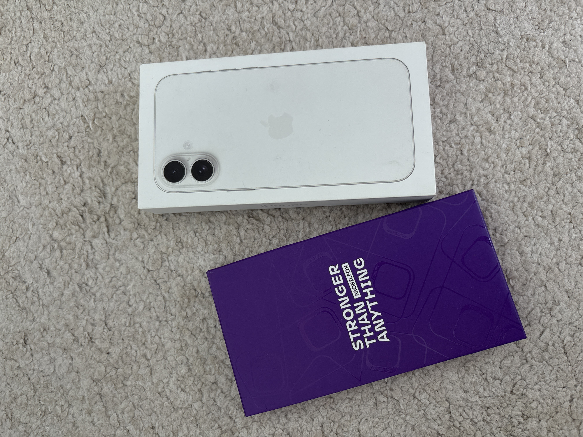 The image displays a flat lay composition of two packaged items resting on a light-colored, shaggy carpet in the city of Sofia, Bulgaria. The carpet is a pale beige or off-white hue and exhibits visible dark fibers, lint, and dust particles.
On the left, a vertical rectangular box of a vibrant purple color is present. The box features an embossed or printed pattern consisting of subtle rounded rectangular shapes. Centered on the box, in white capital letters, is the text "STRONGER THAN" followed by "MOBILFOX" in a smaller font, horizontally centered above "ANYTHING." The word "ANYTHING" is also in white capital letters.
On the right, a white rectangular retail packaging box for a smartphone is positioned vertically. The top surface of this box displays a photorealistic depiction of a white smartphone, likely an iPhone model, viewed from its rear. The depicted phone features a dual camera system with two prominent black lenses arranged diagonally within a raised square module. A smaller sensor or flash component is also visible adjacent to the lenses. The Apple logo is faintly visible on the center back of the depicted smartphone. The white box appears to be in new condition, possibly shrink-wrapped, with minimal surface marks or smudges.
The lighting is even and diffused, without strong shadows, suggesting an indoor setting. No individuals or active interactions are present in the scene.