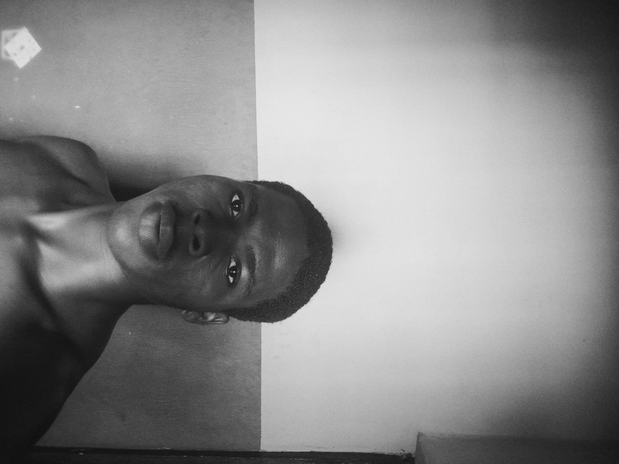 This monochromatic portrait captures a young Black male, shirtless, lying on his back and looking directly into the camera. His head is turned towards the viewer, maintaining a calm, direct gaze.
The image is composed with a distinct background division: a darker, textured surface occupies the left side of the frame, providing a resting place for his head and shoulder, while a contrasting lighter, smoother wall fills the right side. A dark, thin horizontal line at the bottom suggests the edge of a floor or piece of furniture. A small, light-colored, geometric shape (possibly a sticker or reflection) is visible on the darker surface in the upper left corner.
The lighting appears soft and even, highlighting the subject's features and the subtle contours of his upper body. The photograph exhibits a pronounced grainy texture, lending it a raw, intimate, and somewhat vintage aesthetic. A subtle vignette darkens the corners, drawing focus to the center. No text is visible in the frame. The setting is an indoor environment, located in Nkpor, Nigeria.