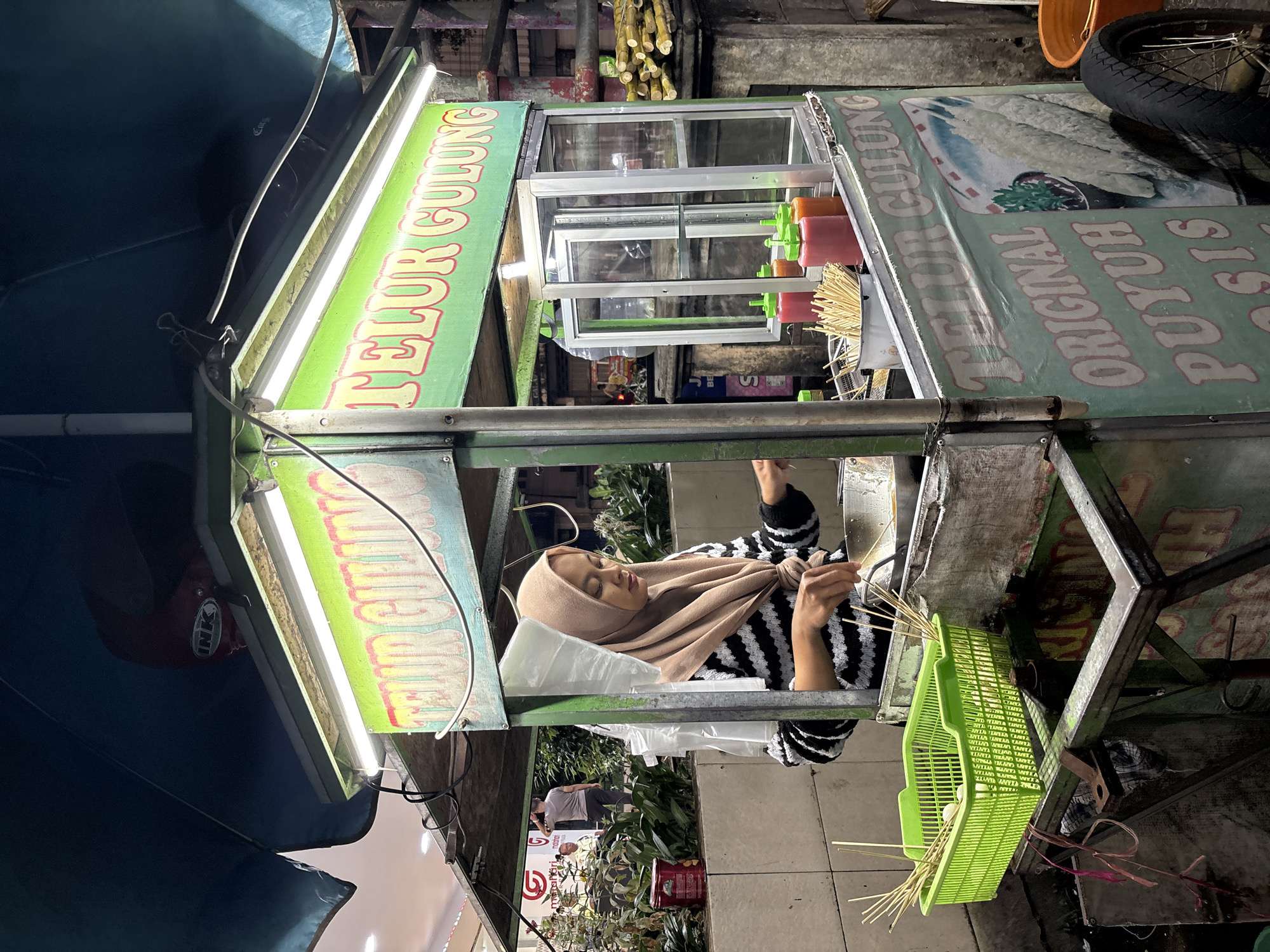 This vertical image depicts a vibrant nighttime street food scene in Jember, Indonesia. The central subject is a woman, wearing a brown hijab and a black-and-white striped long-sleeved top, operating a food stall. She is positioned behind a metal and glass cart, actively engaged in preparing food, holding a stick.
The food cart is well-lit by fluorescent lamps, casting a glow on its green banners. Prominently displayed text on the banners reads "TELUR GULUNG" (Rolled Egg), indicating the specialty sold. Further text includes "ORIGINAL PUYUH PASIS," suggesting variations like quail egg rolled eggs. An illustration of the dish is also visible on one of the banners.
On the cart's counter, clear glass display cases are present, along with several plastic squeeze bottles containing colorful sauces, likely chili sauce and ketchup. A bright green plastic basket overflowing with bamboo skewers is a prominent feature, ready for use in preparing the rolled eggs.
In the background, the scene is dark, characteristic of nighttime, with hints of other street activity, distant lighting, and foliage. Another person is partially visible in the background further down the street. A red helmet with an "INK" logo is discernible hanging on the left side of the stall. The image captures the lively ambiance and practical setup of a typical Indonesian street food vendor at night.