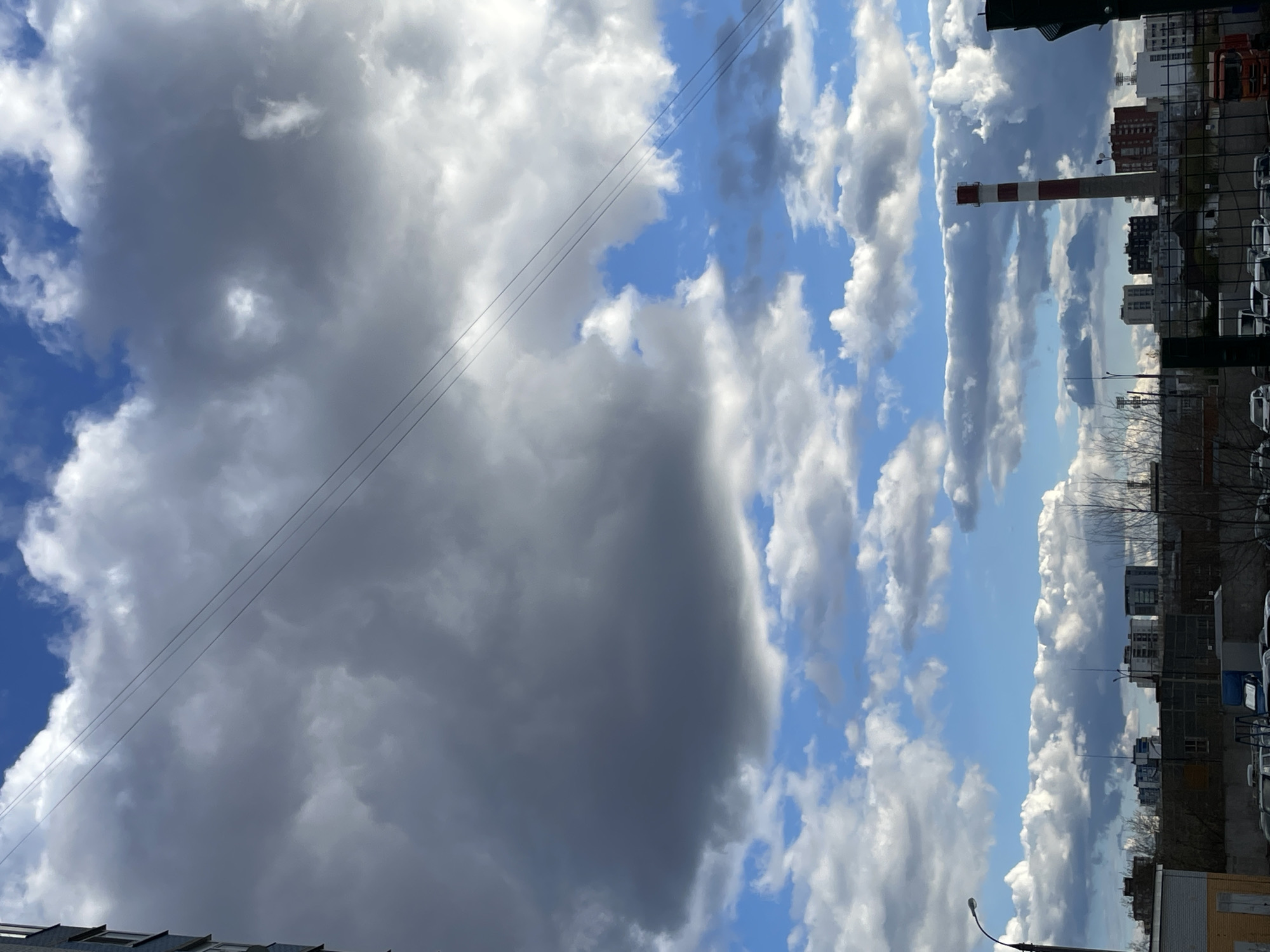 The image captures a dynamic daytime sky over an urban or industrial landscape in Izhevsk, Russia. The sky is a prominent feature, dominated by a large, dark grey cloud mass on the left side of the frame, suggesting heavy cloud cover or an approaching weather system. In stark contrast, the right side reveals a bright blue sky dotted with numerous white and lighter grey cumulus clouds, stretching towards the horizon.
Several thin, dark power lines or wires traverse diagonally across the mid-section of the sky. Below the cloud line and on the right, a cityscape is visible, featuring various buildings, including multi-story residential or commercial structures. A distinct red and white striped industrial chimney rises prominently from this urban setting. Scattered bare tree branches can be seen near the horizon, and some vehicles, likely parked cars, are visible among the buildings. A partial streetlight is present at the bottom right corner, and a fragment of a building's roofline is visible at the bottom left. The scene is static with no specific human activity discernible. The weather appears to be partly cloudy with a mix of sun and heavy clouds. There is no visible text in the image.