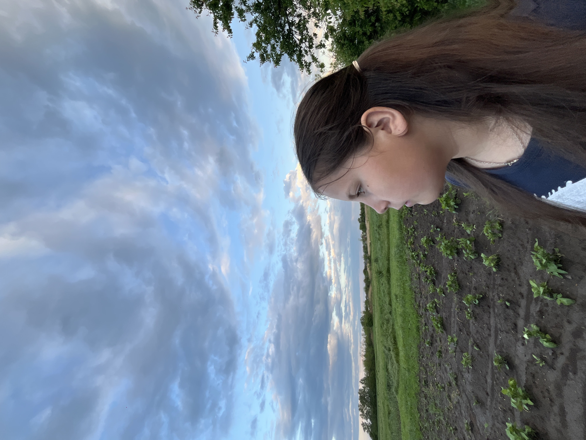 The image presents a landscape with a young woman in the foreground. She has long, dark hair, and is wearing a dark shirt. The location appears to be a field, possibly agricultural, with rows of young plants in the dark soil. The background features a field with green grass and a line of trees. The sky is filled with a mix of clouds, some reflecting sunlight, under a blue sky. The geographical location is specified as Novopokrovskaya, Russia. The angle is such that the field and sky appear slightly rotated in the frame, and the girl appears to be looking down at something out of view.
