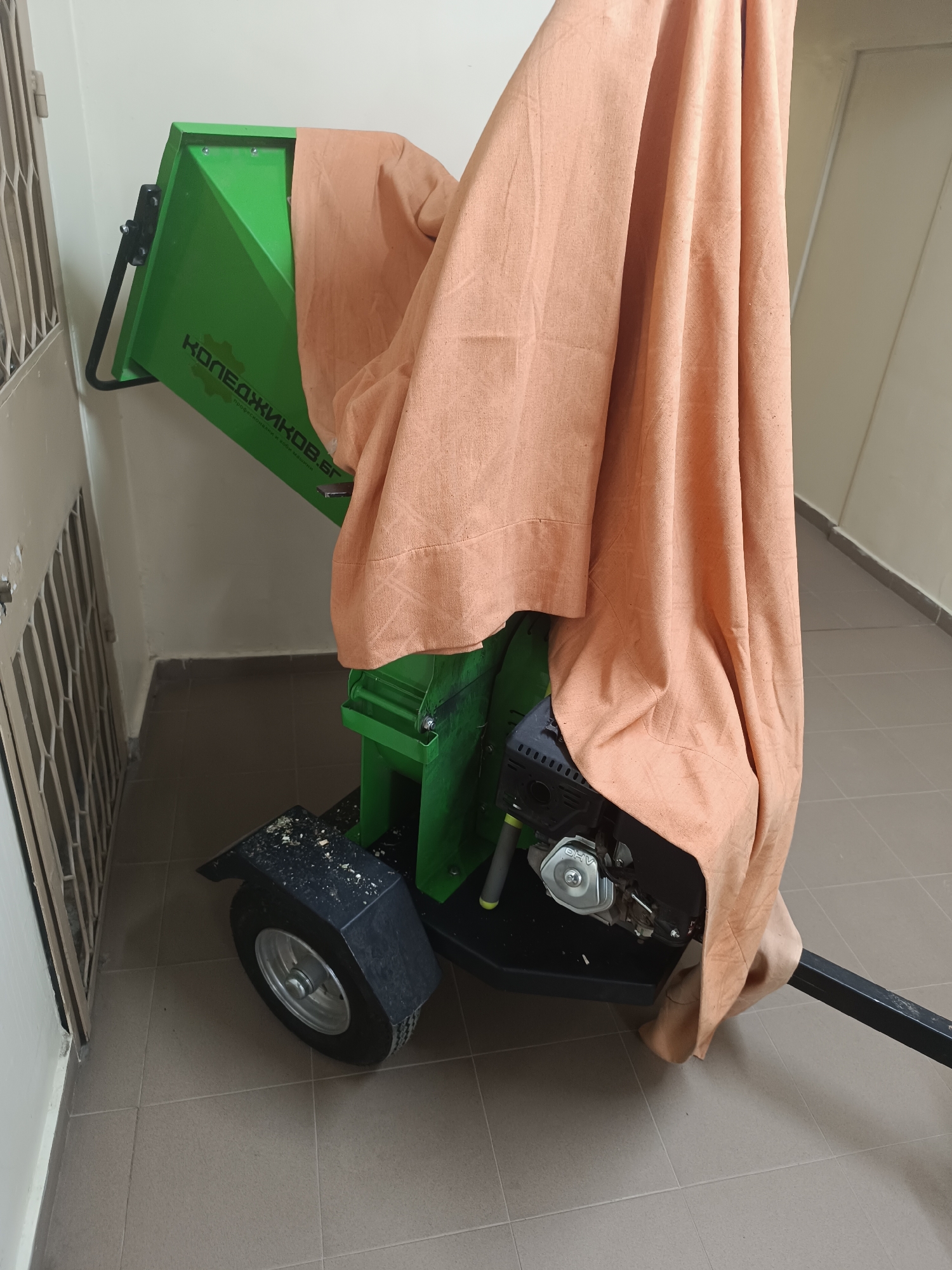 The image shows a green wood chipper partially covered by an orange cloth, parked on a tiled floor indoors, possibly a storage area. The chipper has a black wheel and engine visible. The word "КОПЕАДЖИКОВ.БГ" is visible on the green part of the chipper. The location is the city of Sofia, Bulgaria. The image does not show any people, actions, or interactions. The environment is a simple indoor space with a door to the left and a corridor extending to the right.