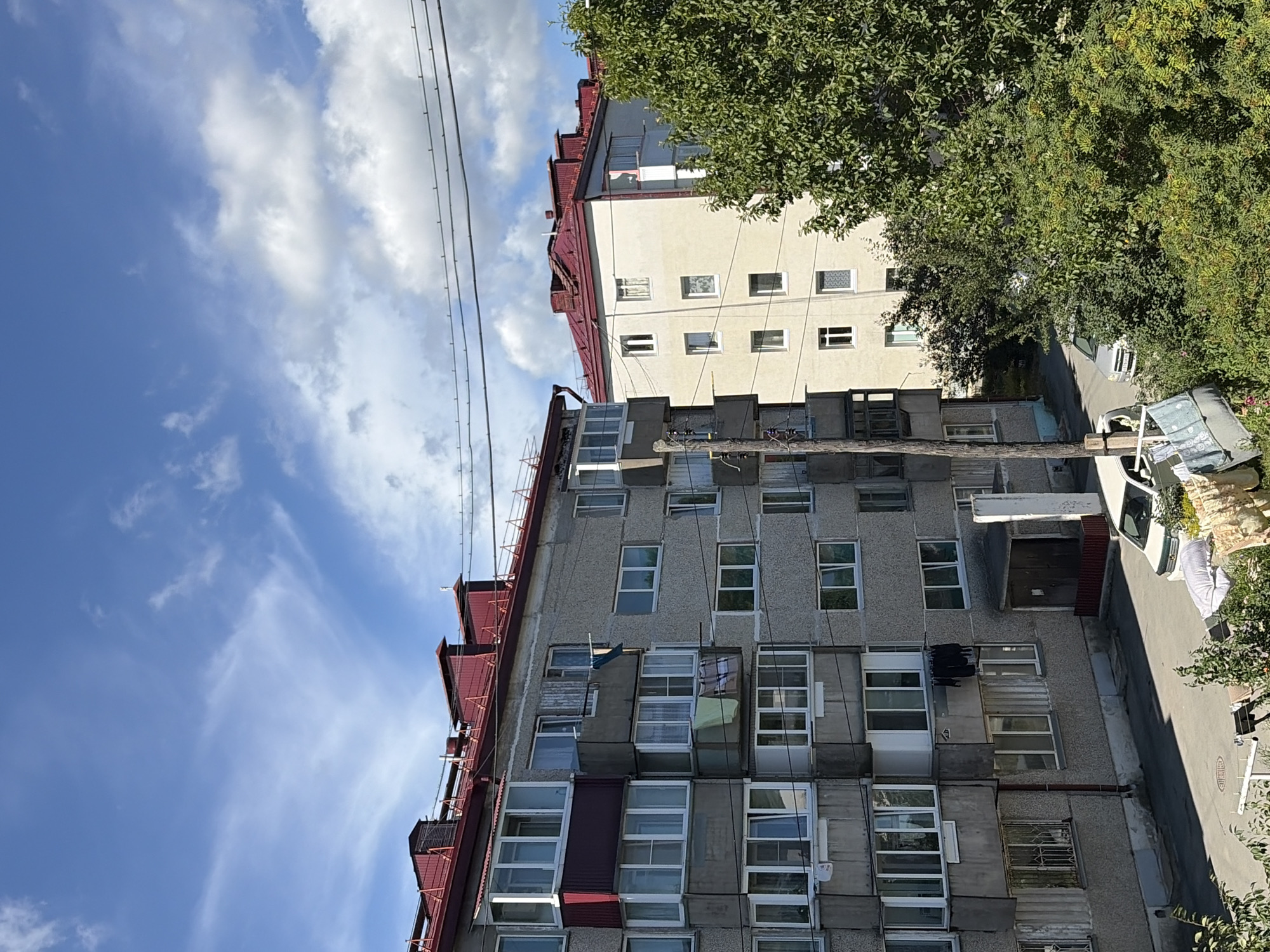 The image captures a street view in the city of Korsakov, Russia, on a bright, cloudy day. Two apartment buildings dominate the scene. The building on the left is older, constructed of concrete, with a red roof and numerous balconies. Laundry is visible hanging from some of the balconies. The building on the right is lighter in color, also with a red roof. In the foreground, a section of a street or parking area is visible, with a car and some outdoor laundry on clotheslines. Lush green trees and bushes flank the buildings and the street. The sky is a vibrant blue, with scattered white clouds. The overall impression is of a residential area in an urban setting.