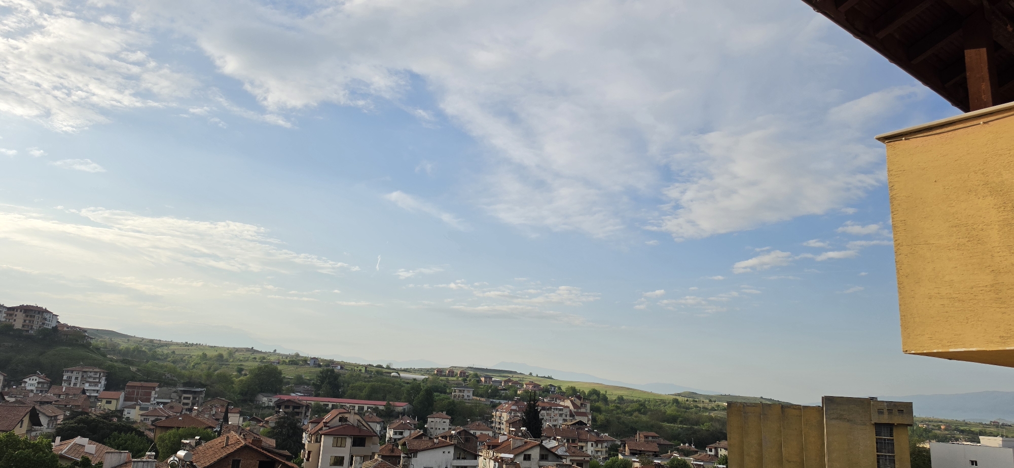 This elevated panoramic view captures a peaceful daytime scene of Sandanski, Bulgaria, nestled within a valley surrounded by rolling green hills and distant mountains. The sky, dominating the upper half of the frame, is a clear, light blue with scattered white, wispy clouds, and a faint aircraft contrail is visible.
The town itself features a dense cluster of residential buildings, many with characteristic red-tiled roofs, extending up the verdant slopes. Interspersed among the houses are larger, multi-story apartment buildings. Lush greenery, including trees and bushes, is abundant throughout the urban landscape and on the surrounding hills. Several rooftops prominently display solar water heaters.
On the far right, a large, textured, mustard-yellow structure, possibly a wall or part of a building's exterior, partially frames the scene. Above it, in the top right corner, a dark brown wooden roof structure with visible beams suggests the photograph was taken from a balcony or an elevated viewpoint. Below the yellow structure, a distinct, light-colored Brutalist-style building with vertical concrete sections and dark windows stands out.
The time of day appears to be late morning or early afternoon, with bright, soft sunlight illuminating the landscape. The weather is fair and clear. No people or specific activities are visible, and there is no discernible text within the image.