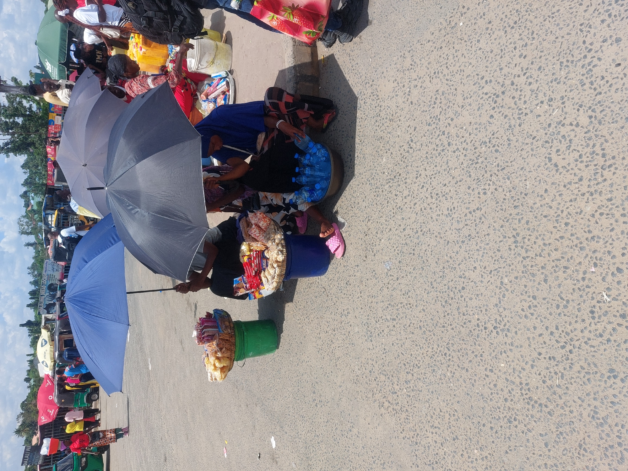 The image shows a street scene in Dar es-Salaam, Tanzania, with multiple vendors selling goods on the side of the road.
**People:** Vendors are seated or squatting on the ground, selling items such as snacks, water bottles, and other merchandise. Some vendors are holding umbrellas to shield themselves from the sun. Some pedestrians are visible in the background.
**Objects:** Umbrellas, buckets, trays, and containers hold various goods for sale, including bottled water, packaged snacks, and other small items. Luggage is piled up next to vendors, suggesting that some may be travelers.
**Actions/Interactions:** Vendors are engaging in sales activities, and it is likely that potential customers are browsing their goods. Some vendors are handling items within their containers, possibly arranging or organizing their merchandise.
**Environment:** The scene takes place on a paved surface next to a road with various vehicles. The sky is visible, with some cloud cover. Trees and other vegetation are also visible in the background, indicating that this street is located within a populated urban area.