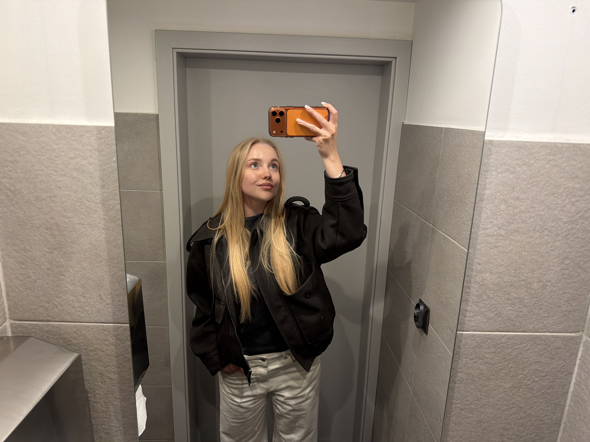 A fair-skinned female with long blonde hair stands in a public restroom, taking a selfie in the mirror with her orange phone. She is wearing a black jacket, a dark shirt, and light-colored pants. The restroom has gray tiled walls and a sink on the left side. The location is in the city of Unknown, Unknown.