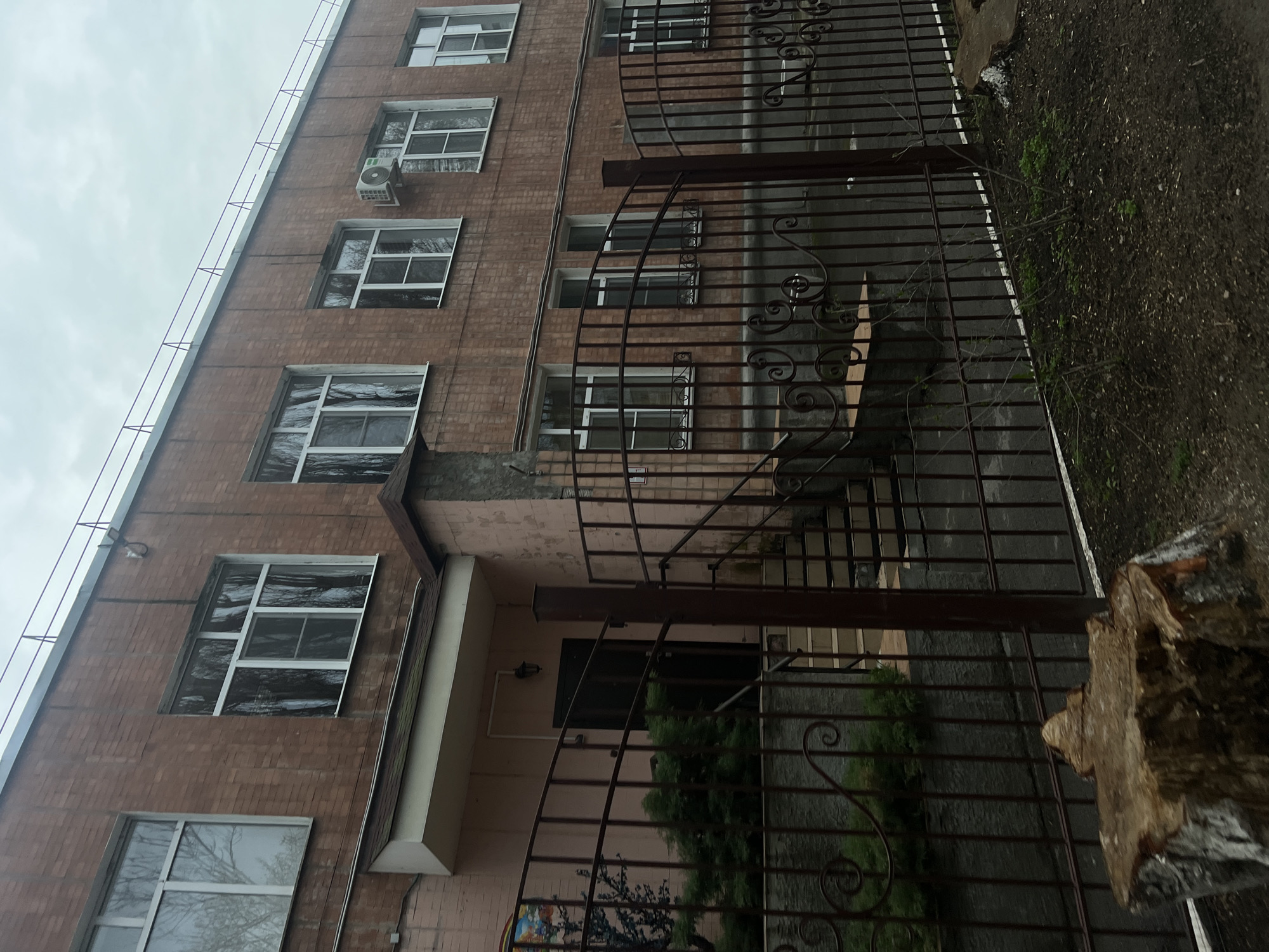 The image shows a brick building with multiple windows, some reflecting the sky, located behind a dark iron fence with decorative elements. An air conditioning unit is mounted on the wall near one of the upper windows. The building has a light pink-painted entrance area with a dark doorway. The environment includes a patch of bare earth and some greenery visible through the fence. A tree stump is visible in the foreground on the right. The image is taken at an angle, appearing to be looking up at the building. It is located in Rostov-on-Don, Russia.