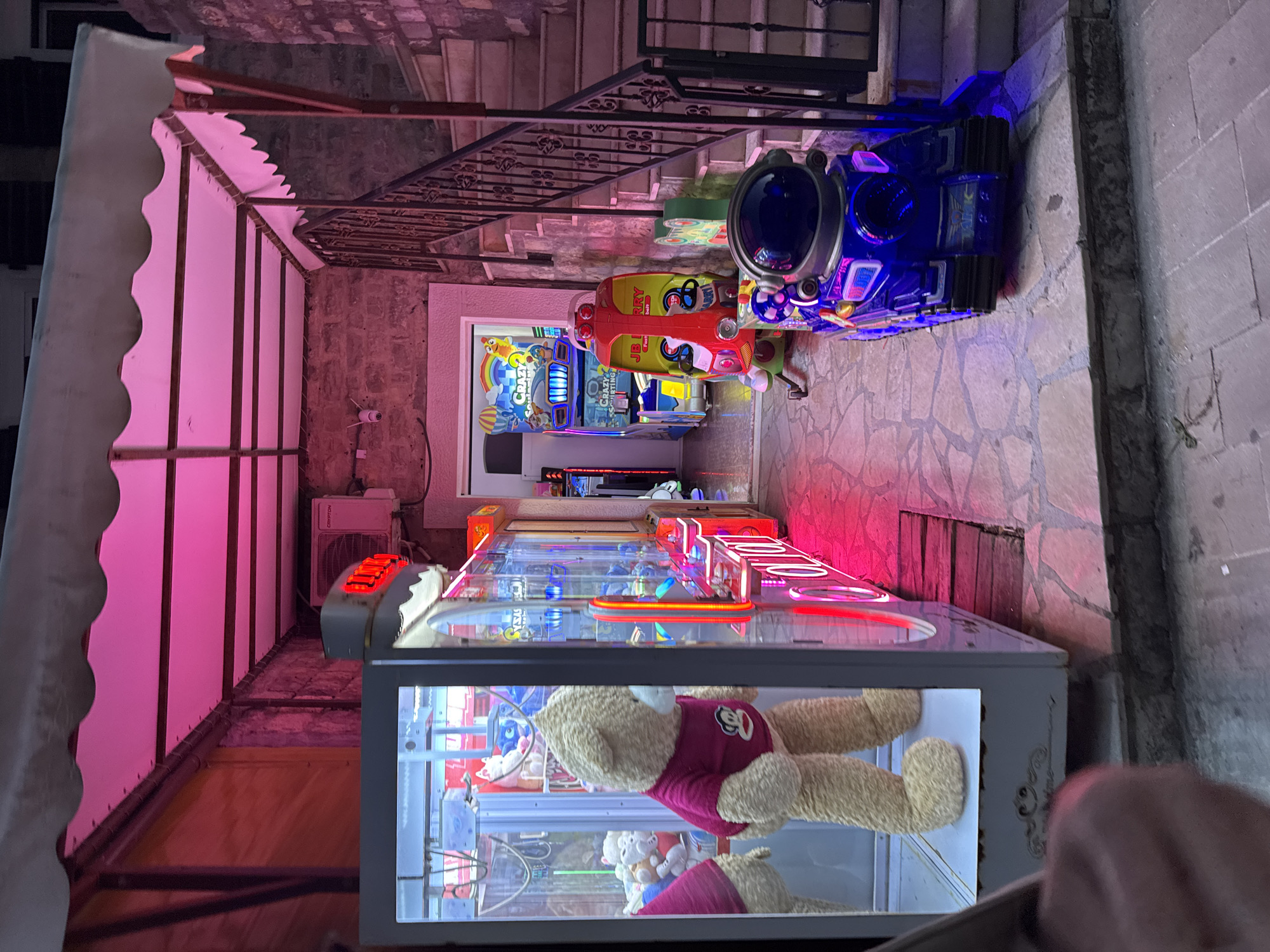 The scene is an outdoor arcade in Kaluđerac, Montenegro. The arcade is located on a stone pathway, which leads up to a set of stairs with wrought iron railings. Along the pathway, there are several arcade machines. The closest machine is a claw crane game filled with stuffed animals. Behind it are additional arcade games, including driving simulators and a "Crazy Taxi" game. An awning, lit with pink lights, covers the left side of the scene. An air conditioning unit is visible on the wall behind the awning. The arcade is surrounded by stone walls. The image also captures a hand holding a device in the bottom right corner.