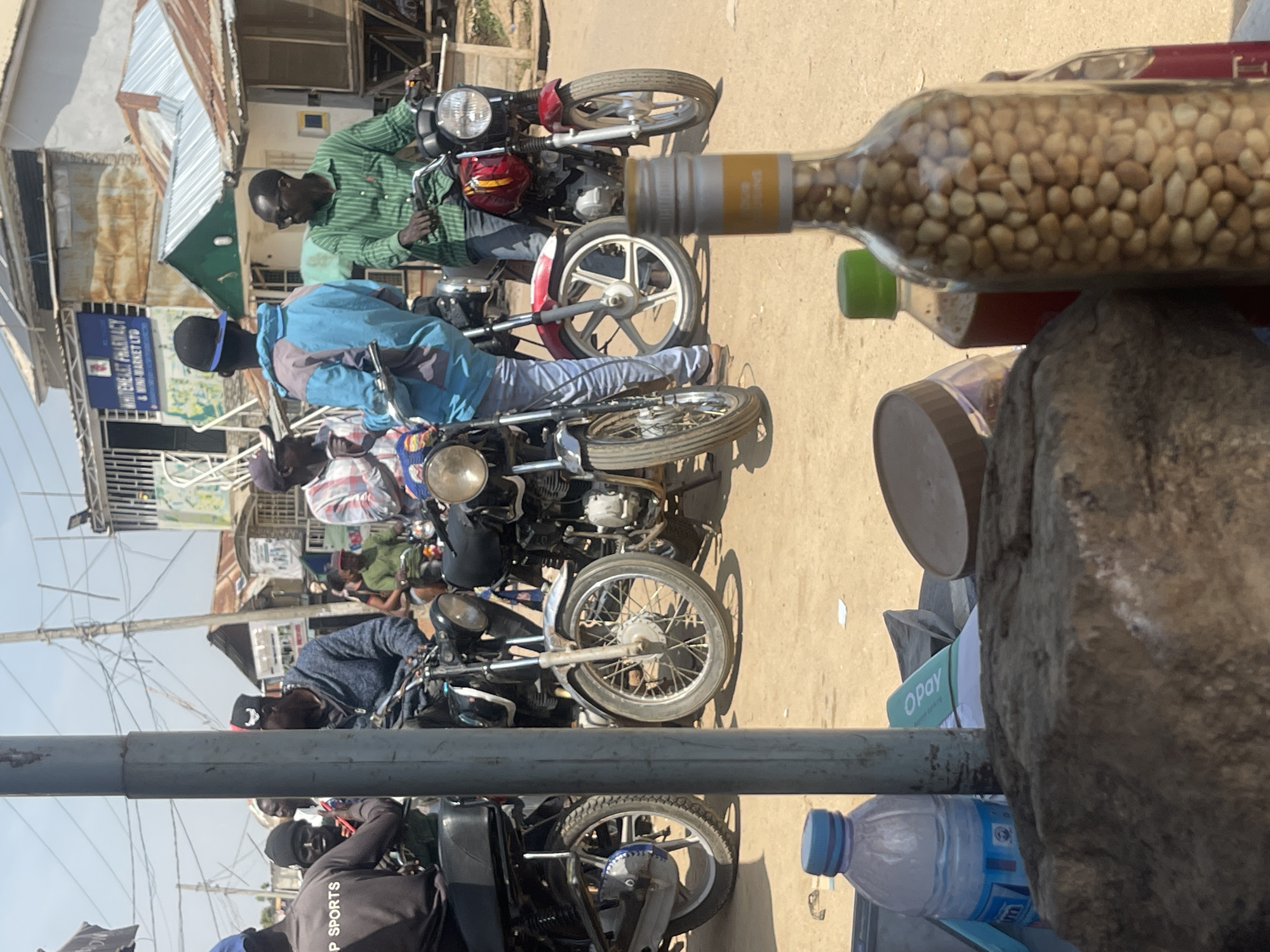 The image depicts a street scene in Abuja, Nigeria, likely a bustling market or a roadside commercial area. In the foreground, a large rock occupies the lower portion of the frame, with a bottle filled with nuts and other items on a surface behind it. A blue plastic water bottle is also visible. The background is dominated by several motorcycles, some parked and others with riders.
A group of men are the primary human subjects, seen on and around the motorcycles. One man in the foreground, partially obscured by a pole, wears a black cap and a blue and grey jacket, and is looking towards the right. Another man, to his right, is wearing a green striped shirt and dark sunglasses, and appears to be seated on a red motorcycle. Further back, other individuals are visible on and around motorcycles, some with casual attire. The environment suggests an outdoor setting with buildings and infrastructure in the distance, including a sign for "WHITEHEART PHARMACY & MINI-MARKET LTD". The ground is unpaved and dusty. The overall atmosphere is one of a lively, everyday scene in a Nigerian urban environment.
