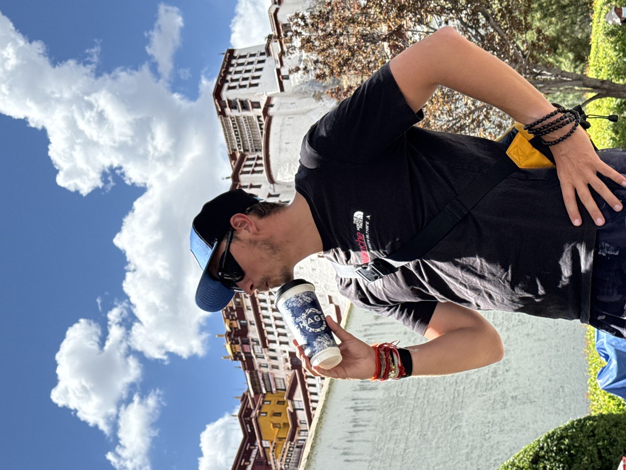 A person is captured in a sunlit outdoor scene, holding a white and blue disposable coffee cup to their lips, appearing to take a sip. The individual is positioned on the right side of the frame, seen from the waist up, and is wearing a black short-sleeved t-shirt, a blue baseball cap, and sunglasses. Their right wrist is adorned with multiple red and orange string bracelets and a wider black one, while their left wrist features several black beaded bracelets. A small yellow pouch with a black strap is visible near their left hip.
In the background, a majestic, multi-story building, characteristic of the Potala Palace, rises prominently. It features distinct white walls, red and yellow sections, and numerous windows, suggesting an ancient and grand architectural style. Lush green foliage and trees are visible around the building and in the foreground. The sky overhead is a vibrant blue, dotted with fluffy white clouds, indicating clear weather.
The activity appears to be a casual moment of refreshment or relaxation, with the person enjoying a beverage amidst significant historical surroundings. The time of day is clearly daytime, with bright sunshine.
Visible text includes a logo on the person's black t-shirt which reads "THE NORTH FACE" along with "NEVER STOP EXPLORING" and coordinates "N°39.87106°E" and "N°116.39758°E". On the coffee cup, a circular logo displays the word "AGE" centrally, surrounded by a blue and white decorative pattern. Given the architectural style and the provided location context, the scene is undoubtedly set in Chengguan District, China, with the Potala Palace as a prominent backdrop.