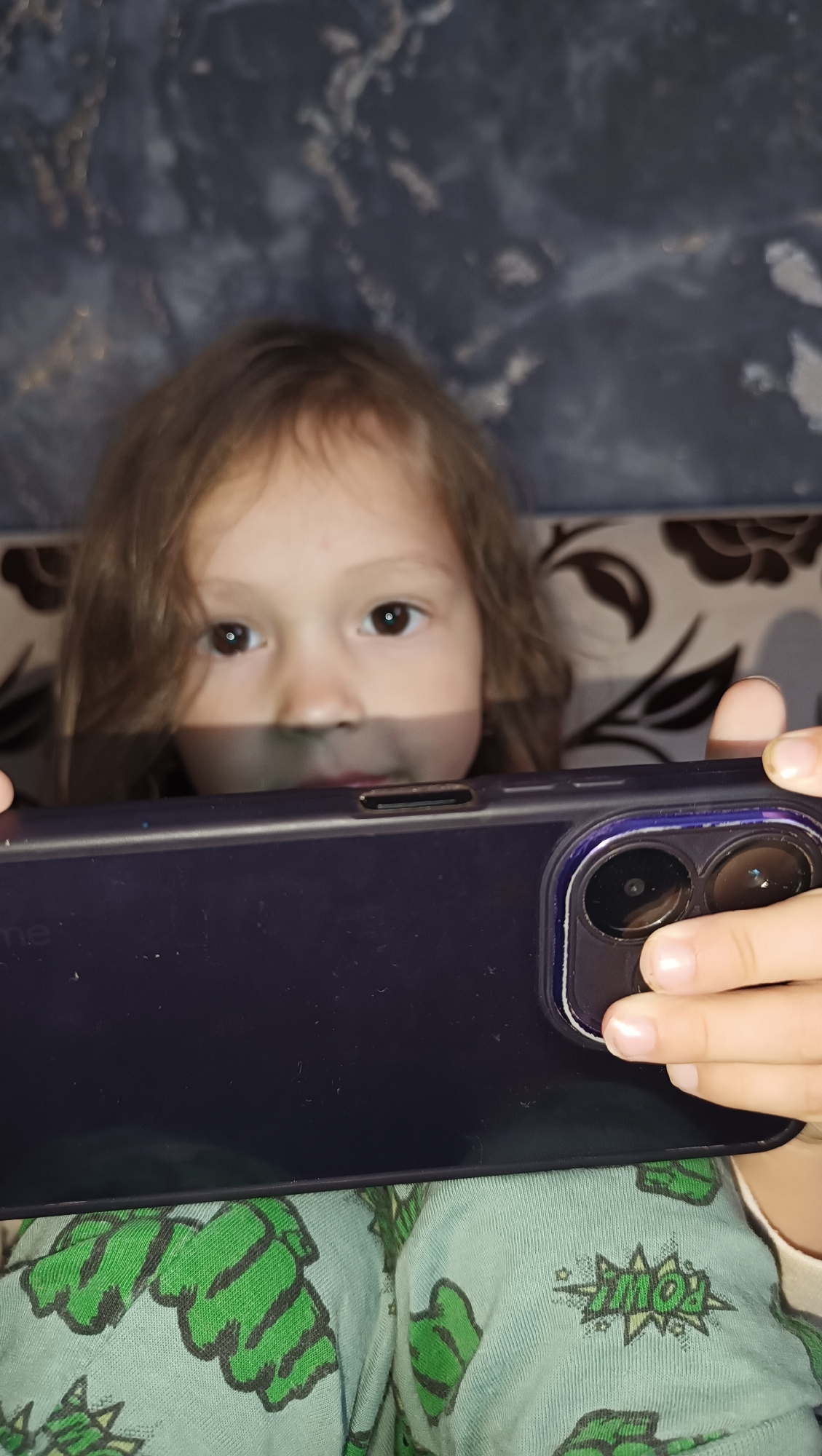 A young girl with light brown hair is holding a dark purple smartphone horizontally in front of her face, obscuring the lower half of her face. She is looking directly at the camera with her brown eyes. Her hands are visible on the right side of the phone, holding it in place. She is wearing light blue pajamas with a green Hulk-themed pattern. The background consists of a dark, textured surface and what appears to be a patterned sofa or cushion. The image is a close-up shot, and no specific location in the city of Karsha, Russia, can be determined from the visual information provided.