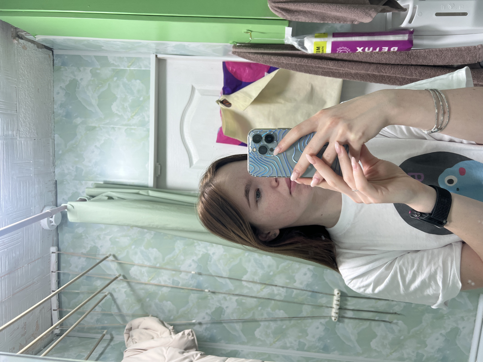 A woman is taking a selfie in a bathroom. She is holding a blue iPhone in her right hand. She is wearing a white t-shirt with a blue cartoon character. She is also wearing a black smartwatch on her left wrist and silver bracelets on her right wrist. The bathroom has light green walls with a textured pattern. A retractable clothes drying rack is visible on the left side of the frame. A brown towel and a package of detox wipes are visible above the woman's head. The location is Chaikovsky, Russia.