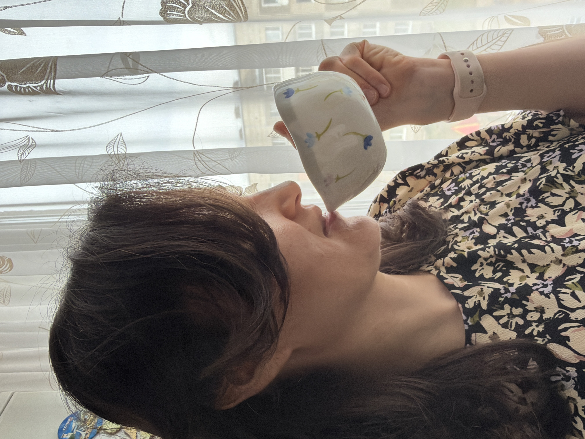 A woman is drinking from a white mug with blue floral patterns. She is wearing a floral dress and a pink smartwatch. The background features a window with blinds and a glimpse of a building exterior. Without additional information, the location in Votkinsk, Russia, cannot be determined.