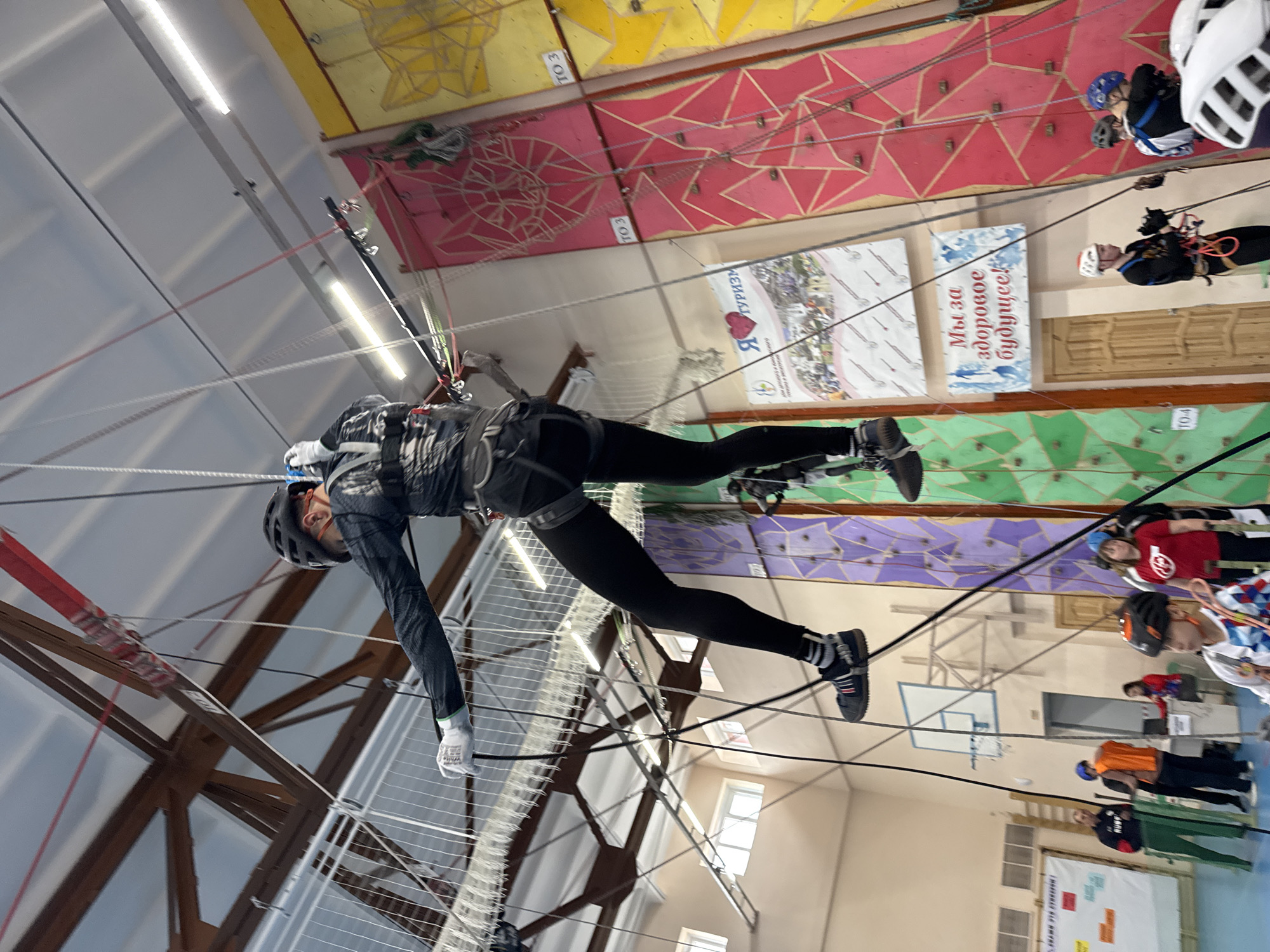 A person wearing a helmet, dark athletic clothing, and gloves is suspended mid-air, attached to a rope course within an indoor structure. The person is wearing a harness, and the cables are attached to it. The background reveals a high-ceilinged interior with a climbing wall decorated with various colors (yellow, red, green, and purple). There are posters on the climbing wall with writing in Russian. There is a basketball hoop mounted on a wall. Several other people stand on the ground, watching. Based on the location of the signs, the image was taken in Orel, Russia.
