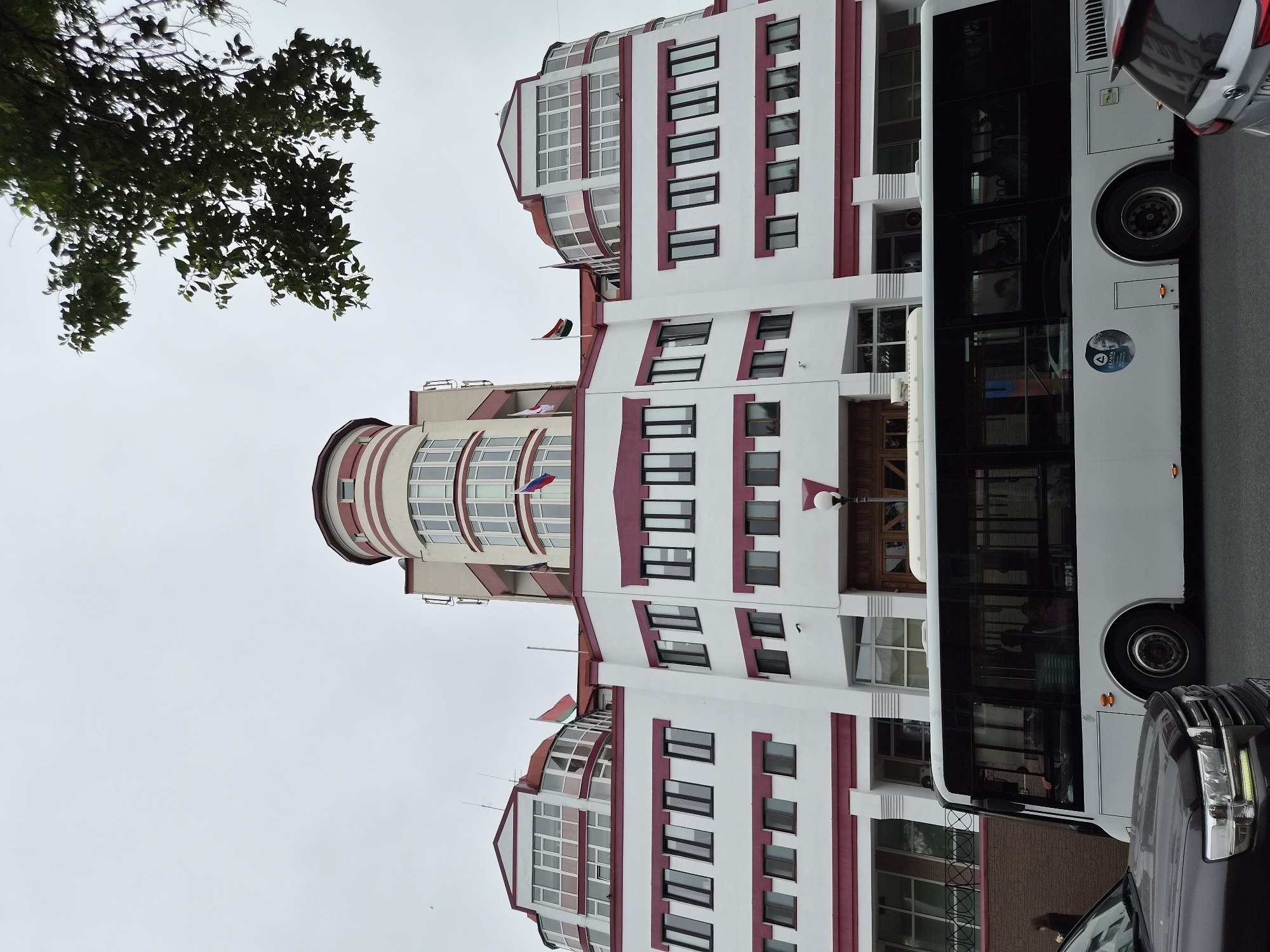 The image shows a multi-story building with a rounded central tower and symmetrical wings, painted white with maroon trim. Several flags are visible on the roof, including the Russian flag. A white bus is driving past the building, with several passengers visible through the tinted windows. A dark-colored SUV is parked in the foreground. The scene appears to be on a street in Vladivostok, Russia, under an overcast sky.