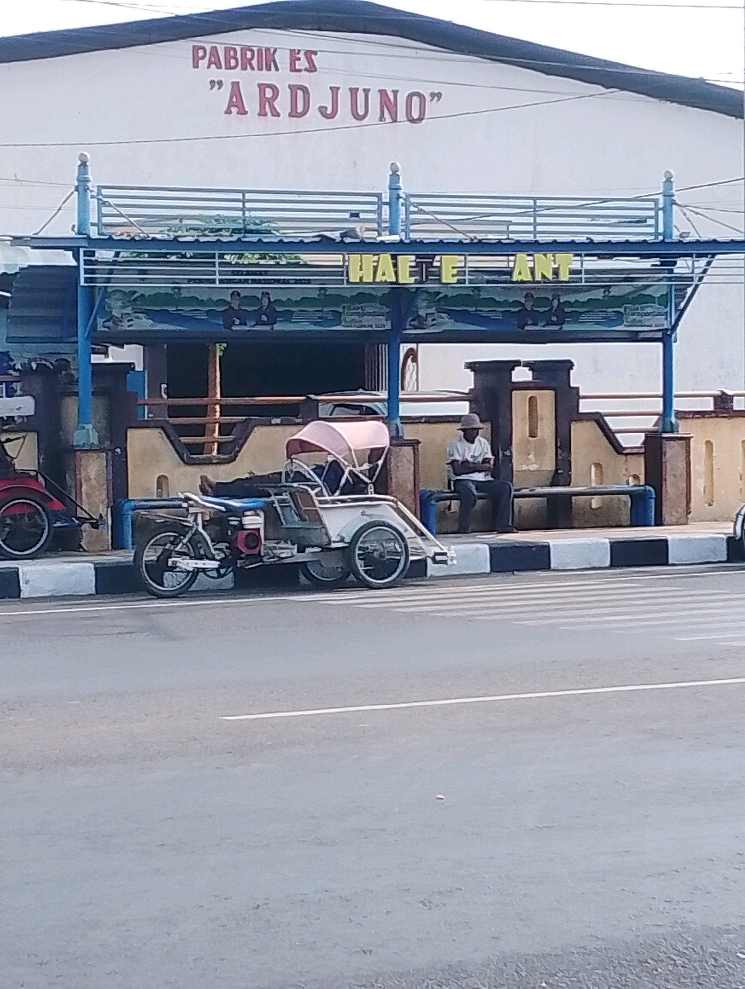 The image captures a street scene in Probolinggo, Indonesia, in front of a building with red lettering that reads "PABRIK ES 'ARDJUÑO'." To the left, a man in a white t-shirt and dark pants, wearing a hat, is sitting on a bench under a covered area. This covered area has a blue railing and a mural depicting people. To the right of the man on the bench, a white motorized becak, a three-wheeled vehicle, is parked. The becak has a pink canopy and a person lying down inside it. Another becak, colored red, is visible further to the left. The scene is bordered by a sidewalk with black and white stripes and a street with visible lane markings. The overall impression is one of a typical street scene in an Indonesian city, likely a commercial area or transport hub.