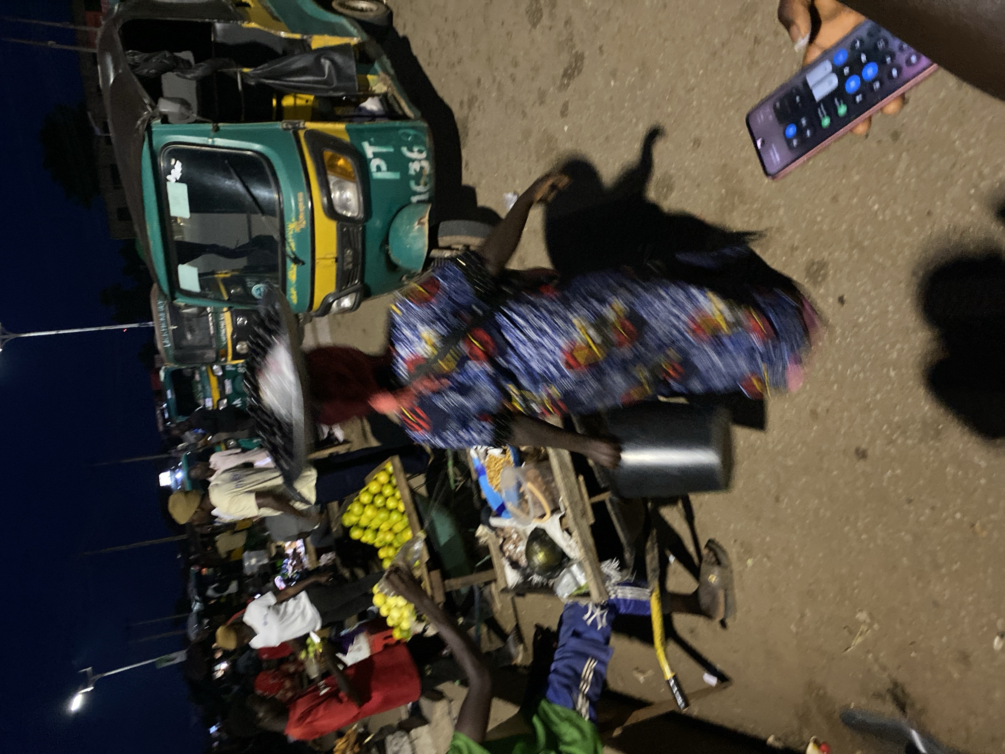 At night in Bukuru, Nigeria, a group of people are gathered around a street market. In the foreground, a person in a blue and red patterned dress walks away from the camera, carrying a large, flat object on their head. To the left, a person in a red t-shirt and dark pants stands near a stall laden with yellow fruits. To the right of the person in the red t-shirt, another person in a white t-shirt and dark pants is visible. Behind them, a green and yellow auto-rickshaw is parked. The ground is unpaved and appears to be dirt or gravel. The sky is dark, indicating it is nighttime. A street light is visible in the upper left corner of the image.