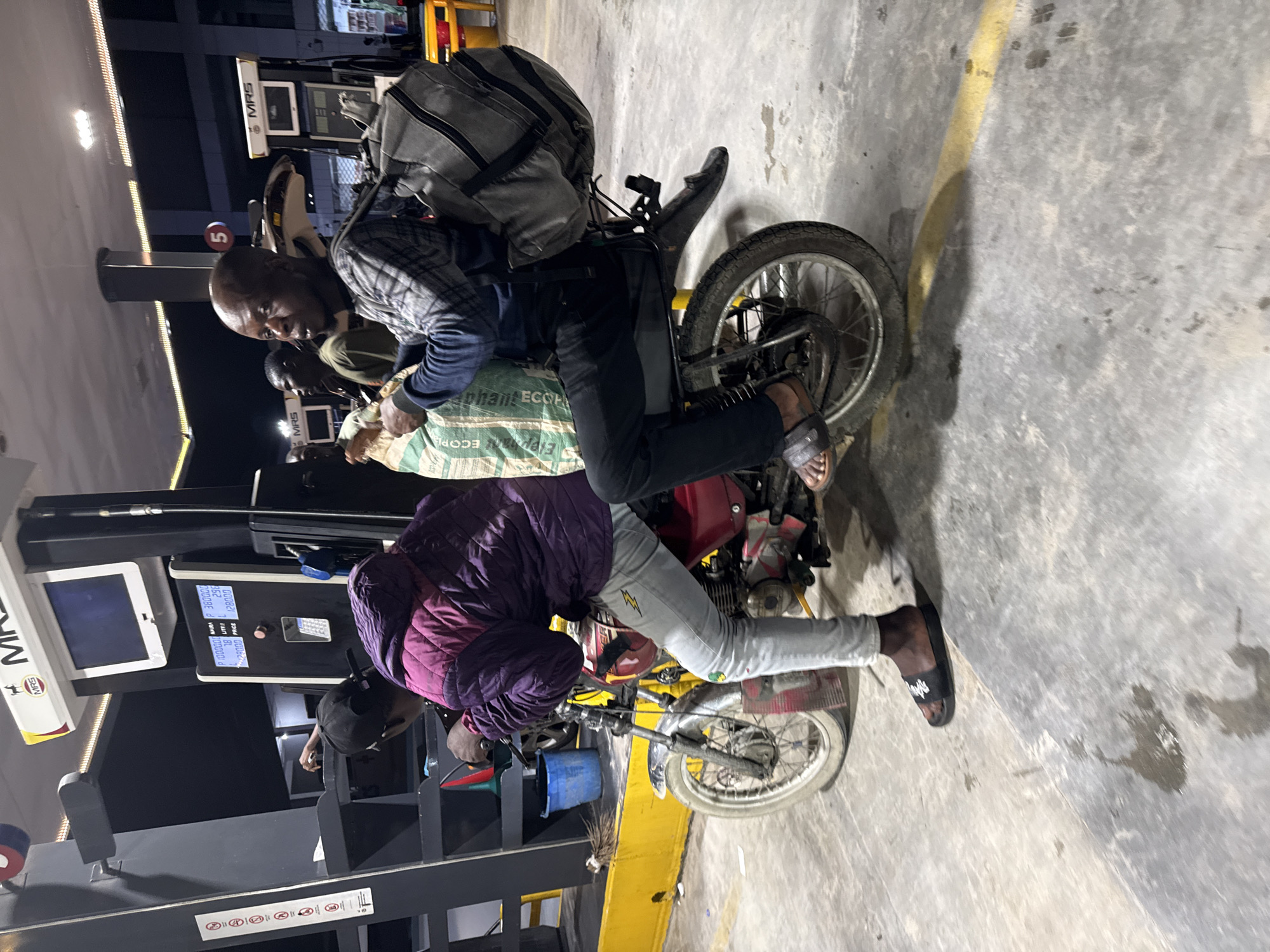At night, an MRS gas station in Ado, Nigeria, is illuminated by overhead lights. A red motorcycle is stationary next to a gas pump. The driver, clad in a purple quilted jacket, light grey jeans, a black cap, and black slides, sits on the motorcycle, gripping the handlebars, with his left foot on the concrete ground. Behind him, a passenger with a shaved head and beard wears a plaid shirt over a dark long-sleeved shirt, dark pants, and dark slides. This passenger carries a grey duffel bag on his back and holds a large, light-colored sack with green text (partially visible as "ECO") in his lap, looking towards the right side of the frame. The adjacent gas pump displays "P 1000000 NAIRA", "78 LITRE", and "128000 PRICE". Additional gas pumps and at least two other individuals are visible in the background. The concrete ground exhibits dark stains and a yellow painted line. A blue bucket and a broom are situated near the pump, and a pole with multiple warning signs is present to the left.