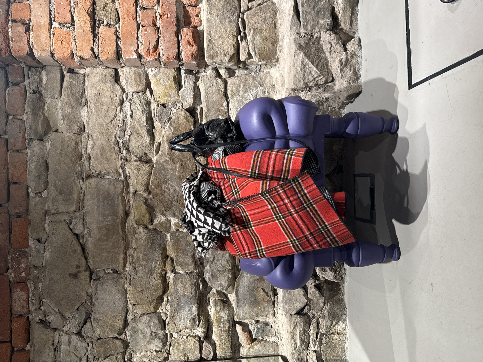 This image depicts an interior scene, presumably within a commercial or renovated historical space in Sofia, Bulgaria. A unique, four-legged, matte purple furniture piece, characterized by its sculpted, multi-lobed form, stands centrally on a light grey, scuffed floor. Draped over the purple object are several textile items: a prominent red plaid fabric with yellow, black, and white stripes covers the front, beneath which a black and white patterned fabric is partially visible. A small black handbag with textured material and braided handles rests on top of the draped textiles. The background comprises a substantial stone wall constructed from irregularly shaped grey and brown stones, with a section of red brickwork visible above and to the right, forming an internal corner. The grey floor features visible scratches and a dark rectangular outline towards the lower right. Distinct shadows are cast from the furniture and objects onto the floor and the lower wall, consistent with internal lighting. No individuals are present in the scene.