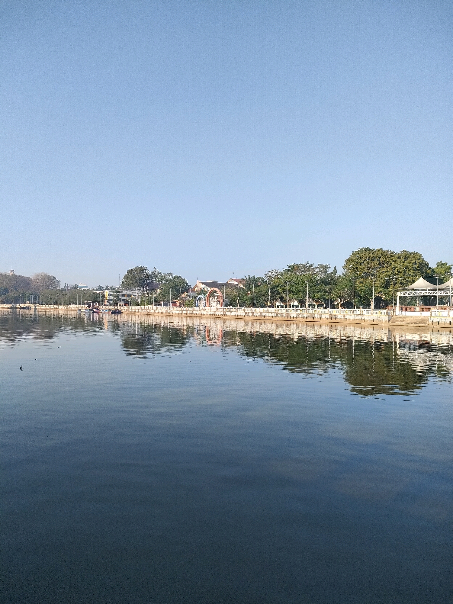 The image depicts a serene waterfront scene in Banjarmasin, Indonesia, likely taken from the vicinity of the Siring Pierre Tendean. The vast expanse of calm water in the foreground reflects the clear blue sky and the structures lining the opposite bank. The bank is adorned with a white railing, interspersed with lampposts, and features a line of trees and buildings. Among the buildings, a white, open-sided structure with a tent-like roof is visible on the right. Further along the bank, a decorative archway with colorful elements stands out. Several boats are moored along the embankment. The water itself is dark and tranquil, with subtle ripples disturbing its surface, and a few dark specks, possibly debris, are scattered across it. The overall impression is one of peacefulness and a well-maintained urban waterfront.
