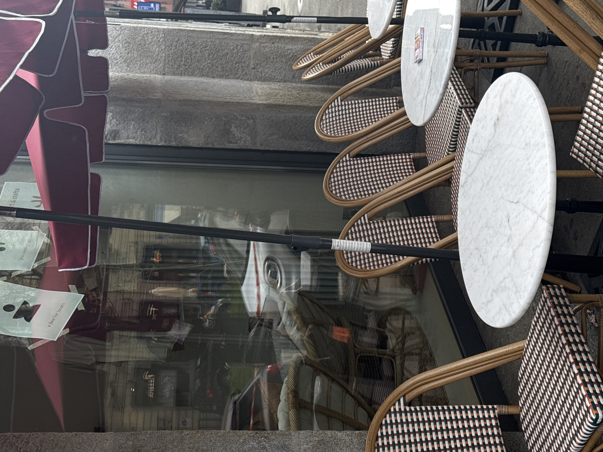 The image depicts an unoccupied outdoor cafe seating area located on a street in Milan, Italy, fronting a building with a textured grey stone facade. Four round tables with white marble tops and dark metal bases are arranged on a dark grey pavement. Eight chairs, crafted from light brown rattan with woven seats and backrests in a repeating checkered pattern of white, dark brown, and dark green, are positioned around the tables. Overhead, two large, partially visible dark red canvas umbrellas with white trim provide shade.
The building's storefront is a large, reflective glass window. Reflections in the window show parked or passing vehicles, including a white car with a horizontal red stripe on its hood, and a darker vehicle. Inside the window, various items, possibly merchandise, are visible. Several white paper signs are taped to the interior of the glass, one prominently displaying the text "a place for success" below a stylized human figure icon. On the marble tabletop furthest right, a small rectangular item, consistent with a pack of cigarettes, is present. No individuals are depicted in the immediate cafe area.