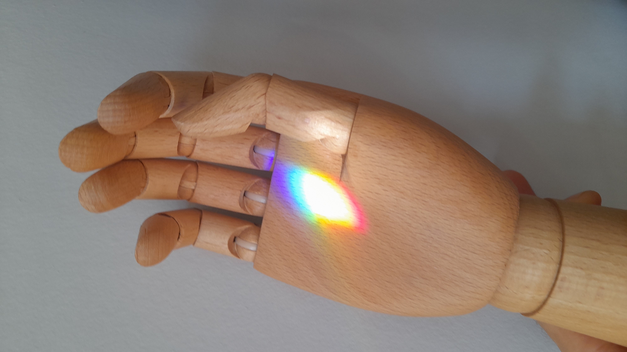 A close-up shot depicts a wooden articulated hand lying on a flat, pale surface. A rainbow-colored oval prism reflection is projected onto the palm of the hand. The fingers of the hand are slightly curled inwards. The color of the reflection ranges from violet through blue, green, yellow, orange, and red. There is no evidence of a location in Saint Petersburg, Russia.
