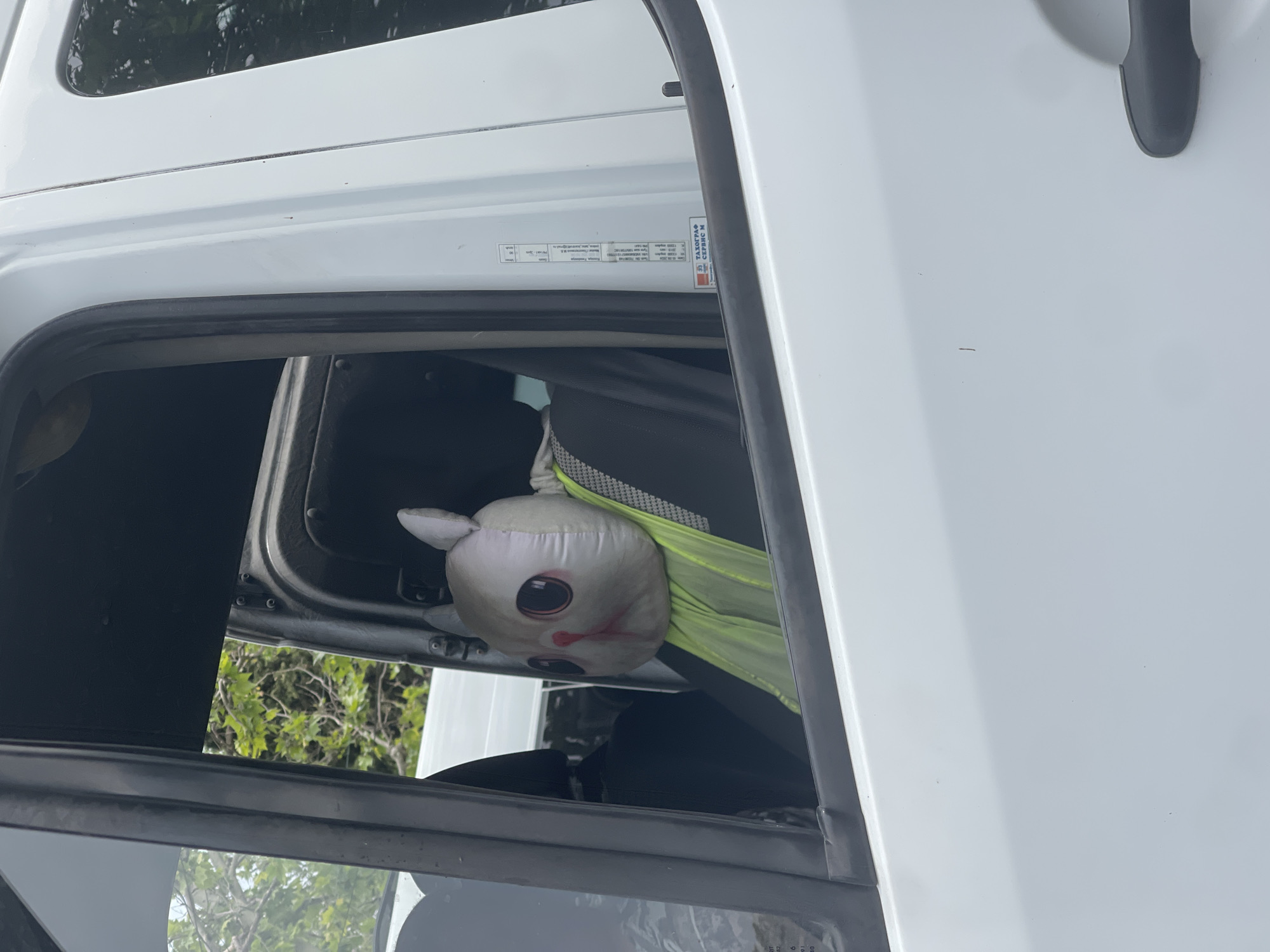 A white commercial vehicle, likely a truck or van, is depicted outdoors in an unknown, unknown city location. The image is taken from the exterior, showing the rear passenger side door. Inside the vehicle, a large, light-colored plush toy resembling a cat or rabbit head with prominent dark eyes and a pink nose/mouth area is seated on the dark fabric rear passenger seat. The plush toy is wearing a bright neon yellow-green safety vest over its torso.
Through the front and rear passenger windows, green foliage from trees and bushes is visible, indicating an outdoor setting. Parts of another white vehicle can be seen in the background through the windows. The white exterior of the depicted vehicle shows minor superficial marks. On the rear door jamb, a white label with Cyrillic text is visible, which includes the words "ТАХОГРАФ СЕРВИС М" (Tachograph Service M) along with other technical specifications and dates. The vehicle's door handle is partially visible on the far right. No human occupants are present in the visible portion of the vehicle.