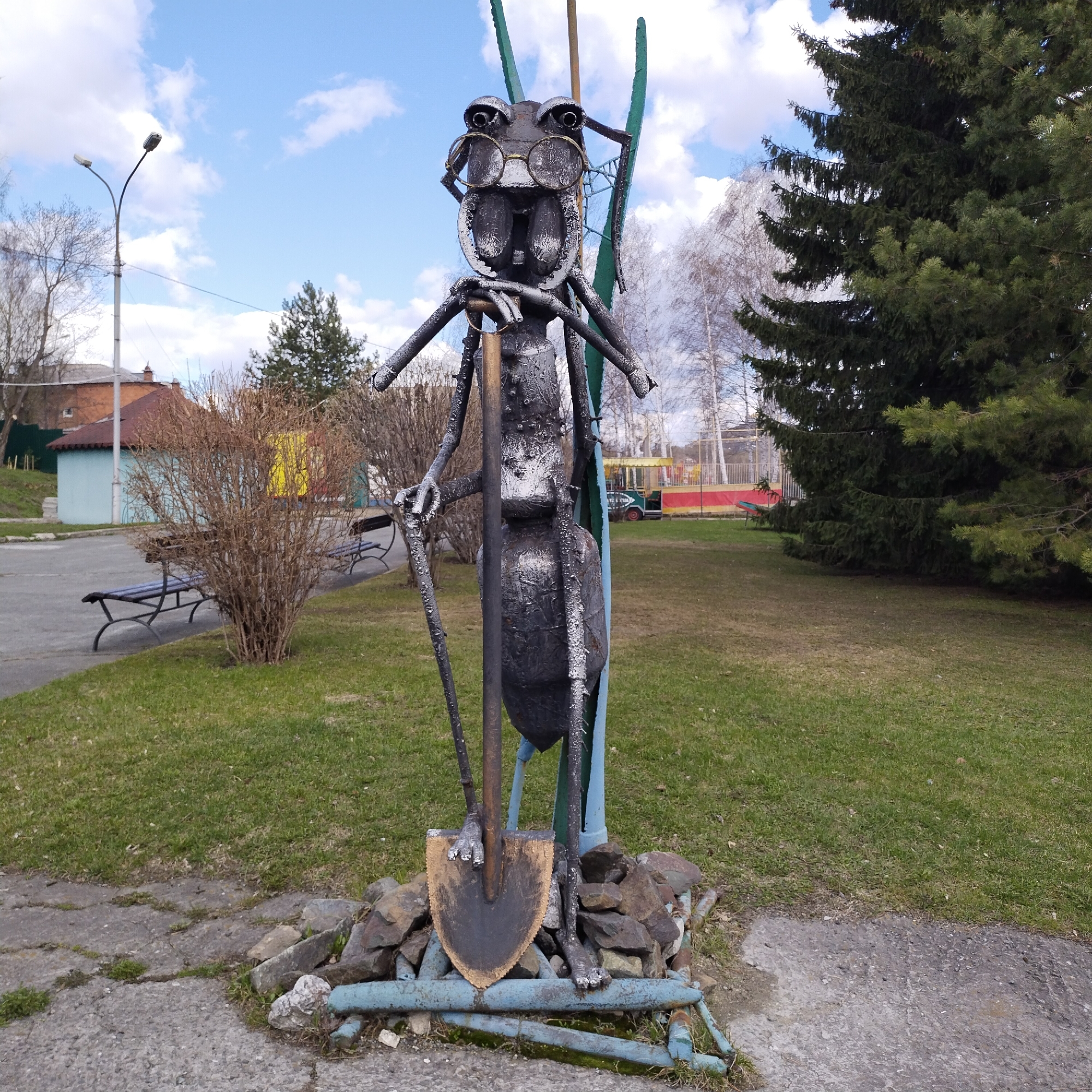 A full shot captures a metal sculpture of an ant wearing eyeglasses and holding a shovel, located in a park-like setting in Nizhny Tagil, Russia. The ant is constructed from metal and painted to resemble an ant, with round eyeglasses positioned on its face. It is standing upright, and a shovel is held by its two front arms, with the shovel blade at its feet. The sculpture is situated on a small base composed of rocks and metal pipes, on a concrete patch. The surroundings are lush green grass, with a large evergreen tree on the right and deciduous trees in the background. In the distance are buildings, including a structure with a red roof and a yellow exterior, possibly part of a playground or park facility. A lamppost is visible on the left side of the frame. The sky is blue with scattered clouds.