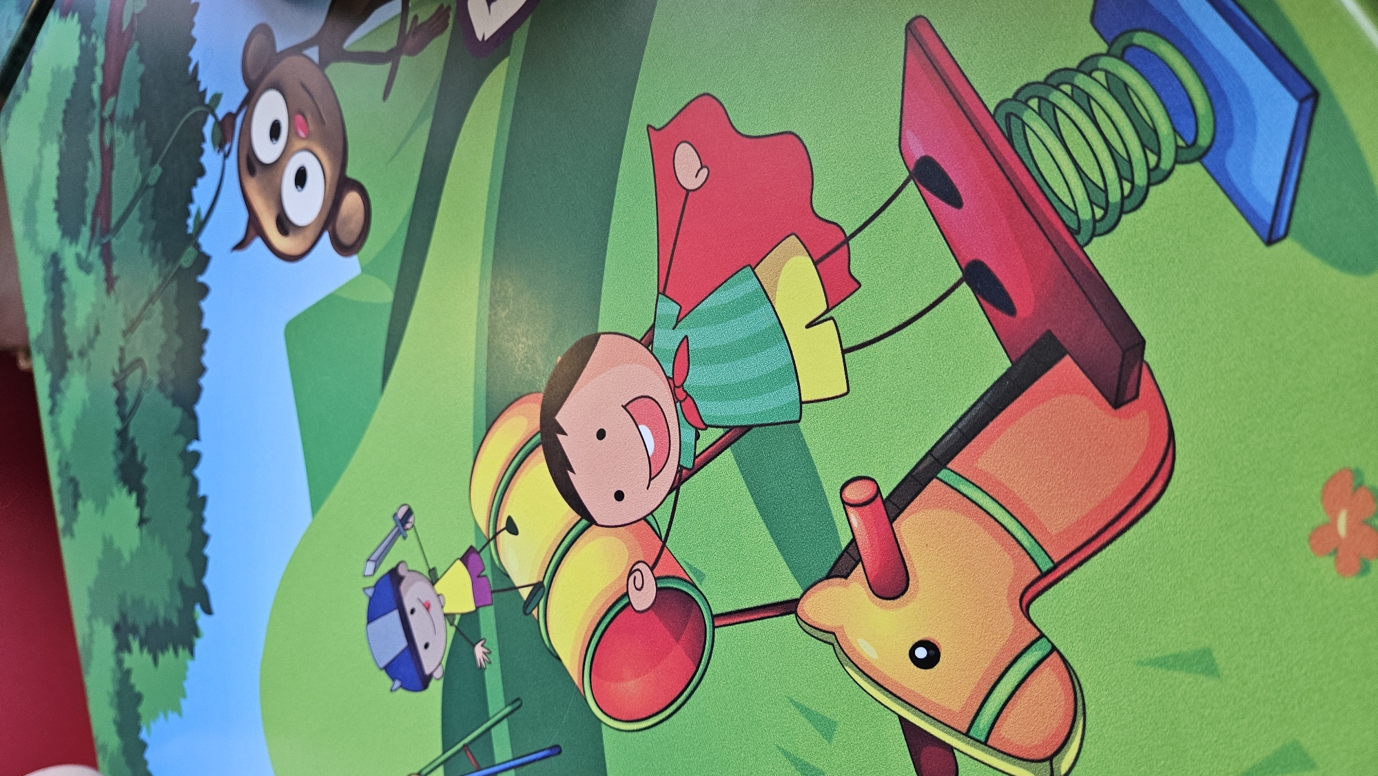 The image depicts a cartoon illustration of a playground scene. In the foreground, a child wearing a green striped shirt, yellow shorts, and a red cape is riding a red spring rider, which resembles a horse. The child has their arms outstretched. Behind them, another child in a yellow shirt, purple shorts, and a blue helmet is standing near a large yellow cylindrical object. Further in the background, a monkey with wide eyes is hanging from a tree branch. The environment is a grassy playground with green hills and a blue sky. There is no discernible location in the city of Bratislava, Slovakia.