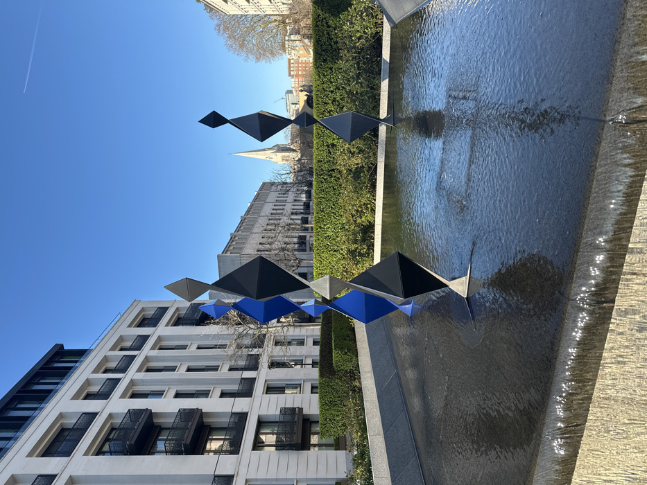 The scene depicts a contemporary urban landscape at Grosvenor Waterside, Gatliff Road, Pimlico, in the City of Westminster, London, United Kingdom, under a clear, bright blue sky.
In the foreground, a dark, shallow rectangular water feature functions as a fountain, with water gently spilling over its front edge. Positioned within this water are multiple geometric sculptures, part of Jonathan Clarke's "The Thread" installation. The leftmost sculpture consists of three vertically stacked rhomboidal prisms, colored royal blue at the bottom, black in the middle, and royal blue at the top. A second, entirely black sculpture of similar design stands further right within the water. A partial view of a third black sculpture is visible on the far right. The water's surface is mildly rippled and reflects the sculptures and the sky.
Beyond the water feature, a well-maintained green hedge separates it from a paved area with several bare deciduous trees. In the midground, to the left, a modern, multi-story residential building with a light-colored facade, dark-framed windows, and black metal balconies rises. To the right, a row of older, light-colored stone buildings with numerous windows is visible. Dominating the central background is the tall, slender, light-colored spire of St Saviour, Pimlico. Further distant buildings are visible to the right. The upper portion of the image is clear blue sky, traversed by a single, thin white aircraft contrail. No people are present in the frame.