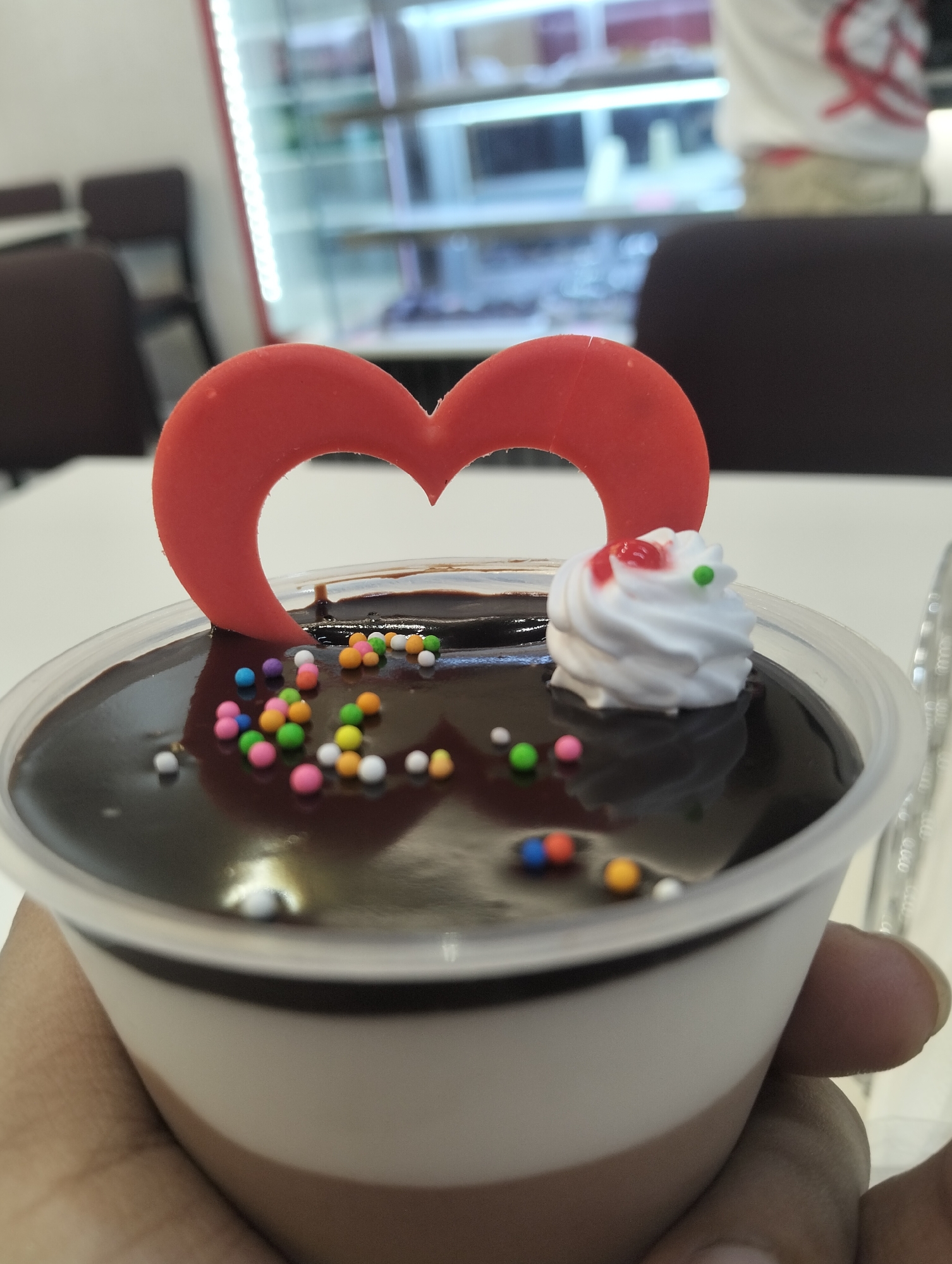 A person holds a clear plastic cup containing a layered dessert. The dessert consists of a bottom layer of brown mousse, a middle layer of light cream, and a top layer of dark chocolate ganache. The dessert is decorated with colorful candy sprinkles, a swirl of white whipped cream with red and green accents, and a red plastic heart-shaped cutout. In the background, a display case with reflective surfaces suggests the location is a bakery or dessert shop. Dark chairs and a white table are also visible. The location is in the city of Dhaka, Bangladesh.