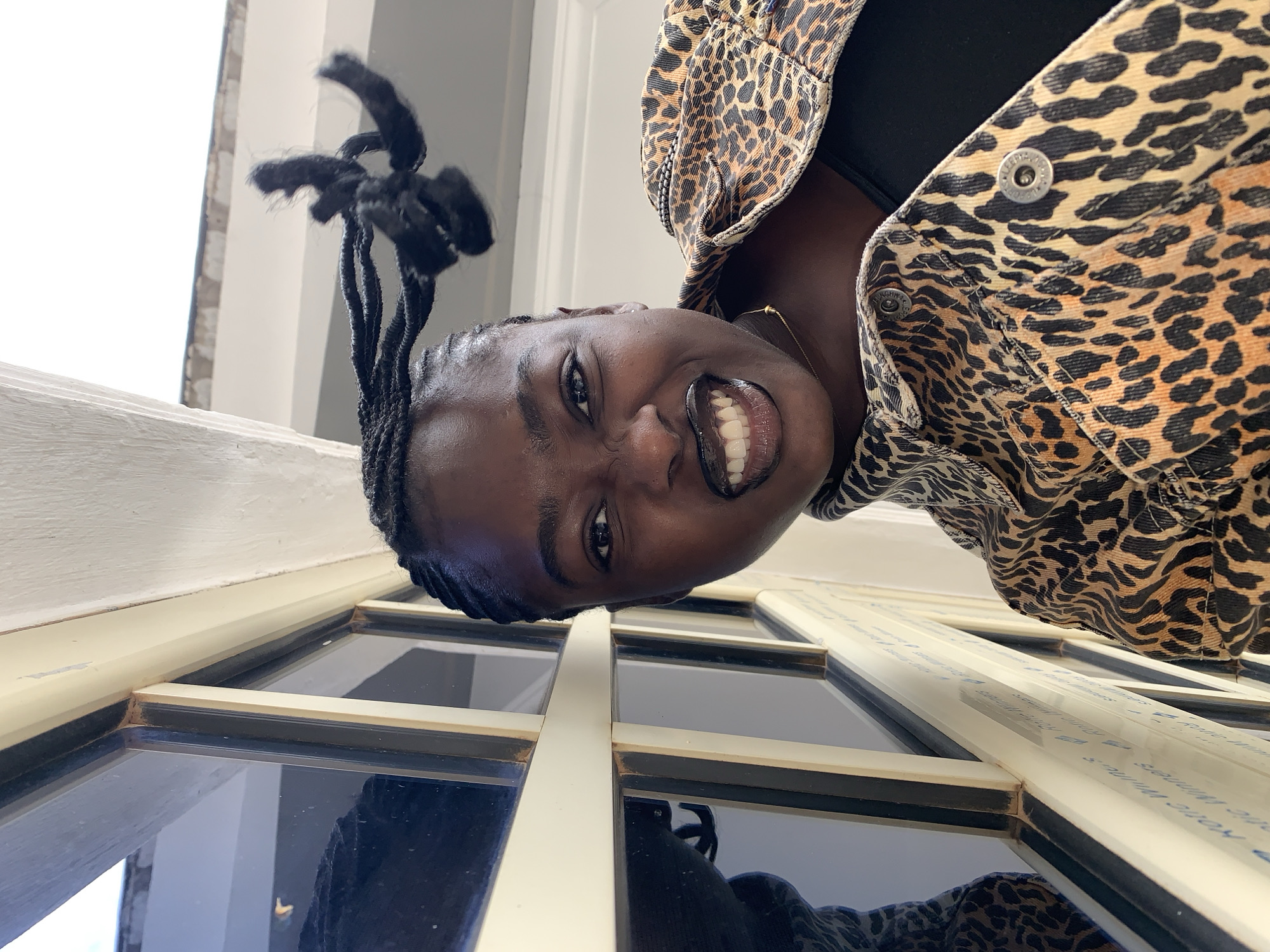 A portrait of a young Black woman wearing a leopard print jacket and a black top, angled with her face towards the camera. She has braided hair and is wearing dark lipstick. The background appears to be a window with a white frame and reflective panes. The image is taken outdoors. The location is Gura Top, Nigeria.