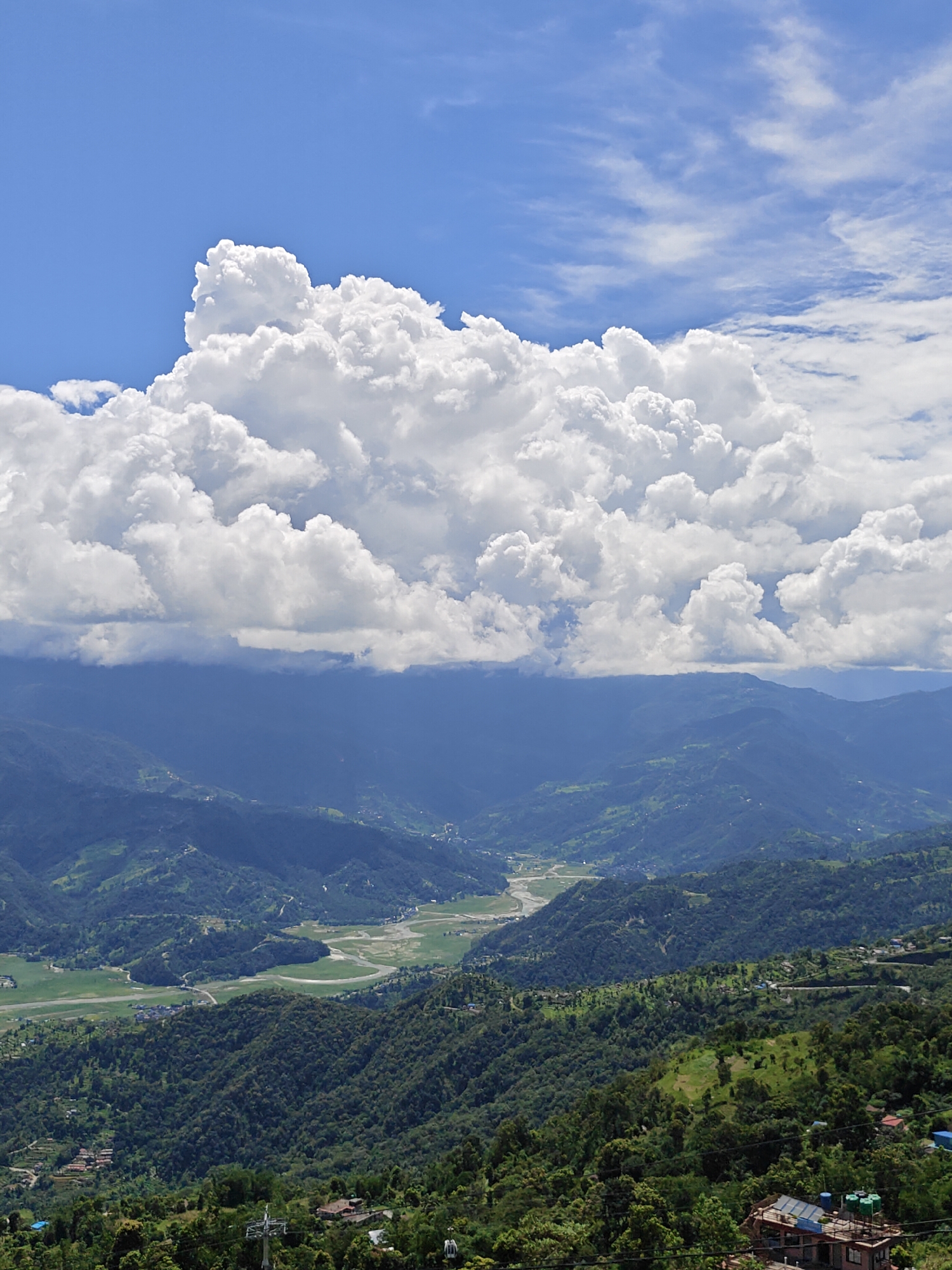 The image captures a scenic vista of the city of Pokhara, Nepal, from a high vantage point. The sky is a vibrant blue, adorned with dramatic, puffy white cumulus clouds that dominate the upper portion of the frame. Below the clouds, a range of verdant, rolling hills stretches across the landscape, diminishing in intensity towards the horizon. The hills are densely covered in lush green forests, with patches of lighter green indicating cultivated fields or clearings. A winding river or a channel of water is visible in the valley between the hills, reflecting the sky and appearing light brown in color. Small settlements with scattered buildings are nestled within the hillsides and along the valley floor. In the foreground, the closer hills are also covered in dense vegetation, with a few structures, including what appears to be a cable car tower and some houses, visible. The overall impression is one of natural beauty and tranquility, with the vast sky and expansive landscape creating a sense of depth and scale.
