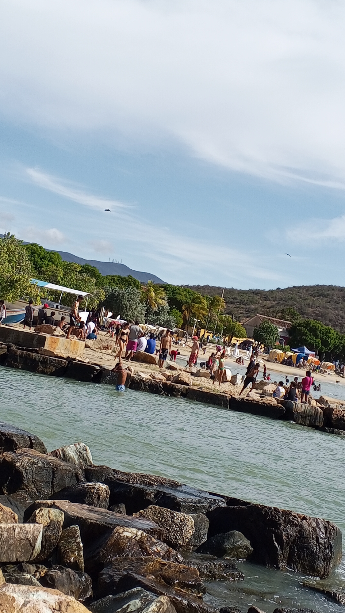 The image depicts a beach scene, possibly in Pedro González, Venezuela, on a sunny day. In the foreground are large, dark rocks bordering the water. The water is a light greenish-blue, and there are gentle ripples on the surface. Beyond the water, a concrete breakwater or pier is lined with numerous people engaged in various activities, such as sitting, standing, and wading in the water. A white vehicle with an open-air seating area is parked along the breakwater. Behind the breakwater is a sandy beach with more people, some under umbrellas. The background includes green trees, hills, and a building or structure, with a slightly hazy blue sky overhead and some clouds. There are two objects flying, likely birds.