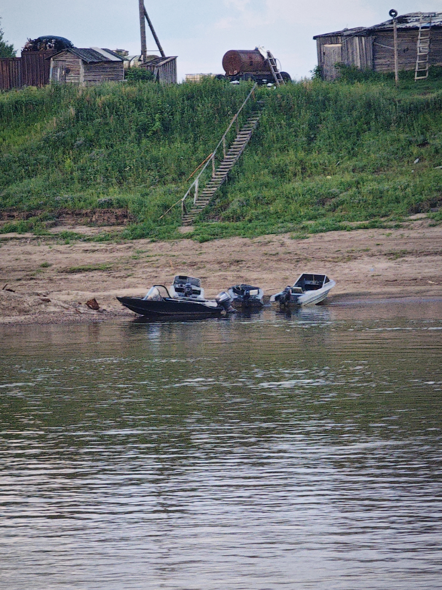 The image depicts a riverbank scene in Vorogovo, Russia. The foreground is dominated by the rippling surface of a body of water, reflecting the green vegetation of the opposite bank. On the sandy shore, three small motorboats are moored. Two of the boats are closer to the viewer, with one partially submerged, while a third boat is further back on the bank.
The middle ground features a steep, green, overgrown embankment leading up to a cluster of rustic wooden structures. A wooden staircase ascends the embankment, providing access to the buildings. Among the structures, a large, cylindrical, rusted tank on wheels is visible, as well as several sheds. The background shows a hazy sky with some faint clouds. The overall impression is one of a remote, possibly abandoned, settlement by a river.