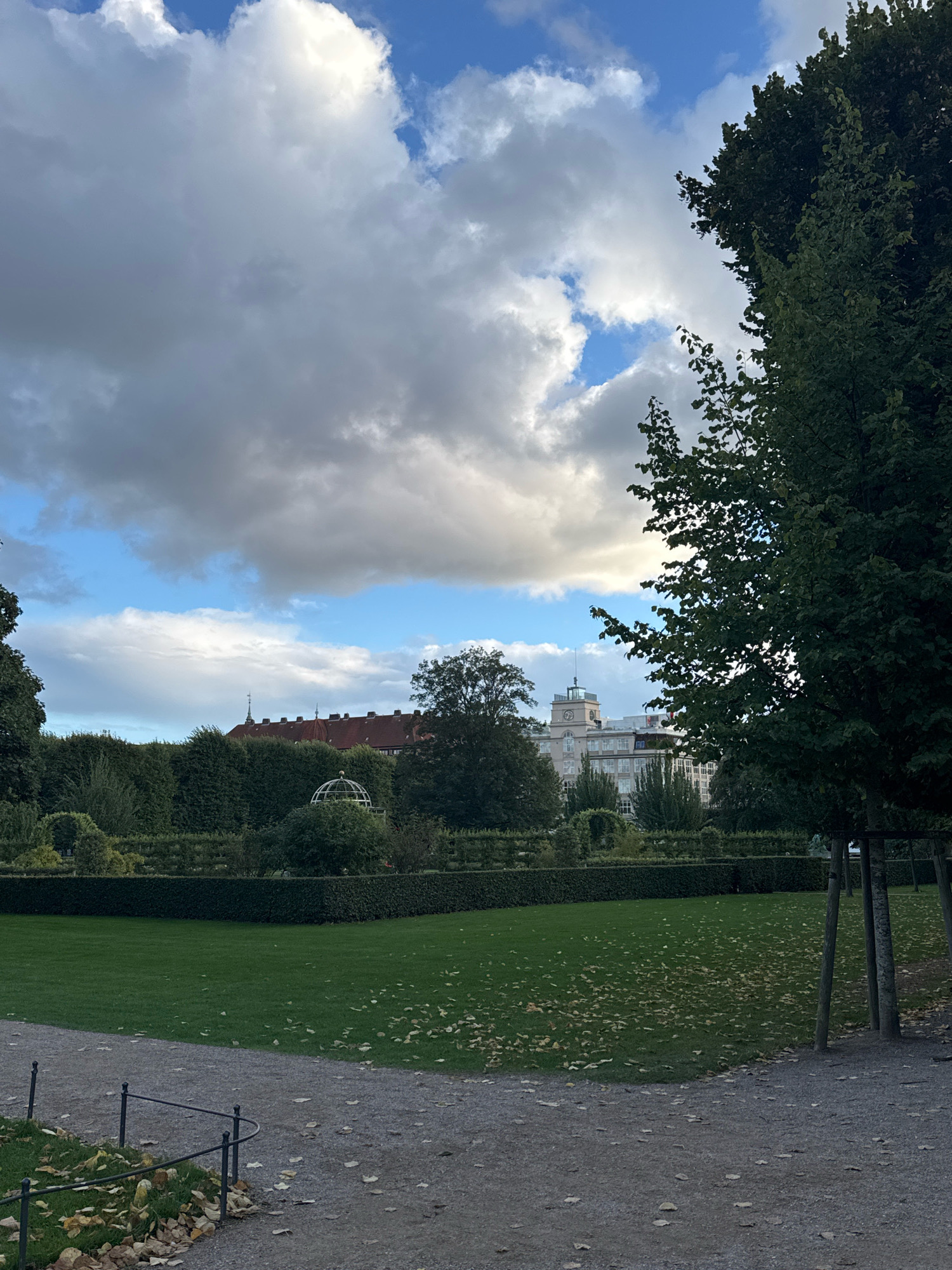 In a park, a gravel path with a low metal fence runs diagonally across the foreground. Beyond the path, a manicured lawn is dotted with fallen leaves. A trimmed hedge forms a dark green boundary for the lawn, and behind it, more trees and shrubs are visible. In the middle ground, a domed structure, possibly a garden folly, is situated within the greenery. In the background, a large building with a light-colored facade and a clock tower stands beneath a dramatic sky filled with large, fluffy clouds. Another building, partially obscured by trees, features a red roof and arched windows. A large, dark green tree dominates the right side of the image, its branches reaching towards the sky.