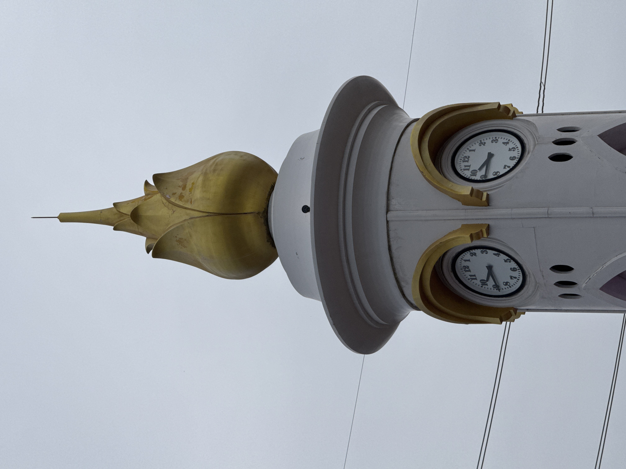 The image shows the top of a clock tower in Chalong, Thailand, against a cloudy sky. The tower is white with gold accents. At the very top is a gold, petal-shaped ornament topped with a thin spire. Below this is a white tiered section, and further down are two clock faces, one on each visible side of the tower. Both clocks have white faces with black numbers and hands, showing approximately 9:45. Below the clocks are three circular openings. The tower appears to be made of concrete or stucco. Overhead, two parallel wires stretch across the frame.