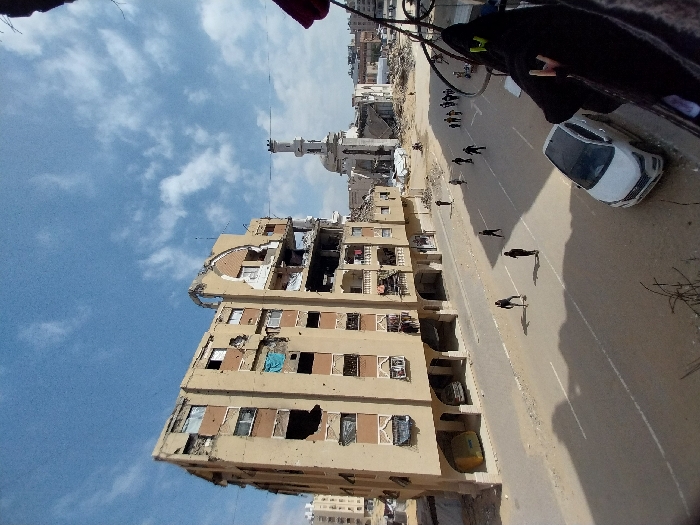A high-angle forensic view captures a street scene in Khan Yunis, Palestinian Territory, under a partly cloudy blue sky. In the immediate foreground, a dark fabric object hangs over a railing on the right, partially obscuring the view. Below, a white SUV (Hyundai Tucson or similar) is parked on the right side of a two-lane asphalt road with faded white markings. Approximately 10-15 individuals, predominantly dressed in dark clothing, walk on the road, casting long shadows to the left, with one individual bending low near the left curb.
To the left of the road stands a multi-story building, heavily damaged with significant structural collapse, exposed interiors, and numerous impact points on its tan-colored facade. Some window openings are covered by blue or other colored tarps. The ground level features arched recesses and a yellow container. Directly across the street, in the midground-background, a mosque with a partially standing, slender minaret exhibits severe damage to its main structure, surrounded by extensive rubble and debris. Beyond the mosque and damaged building, additional structures are visible, many appearing partially or fully destroyed, indicating widespread urban devastation.