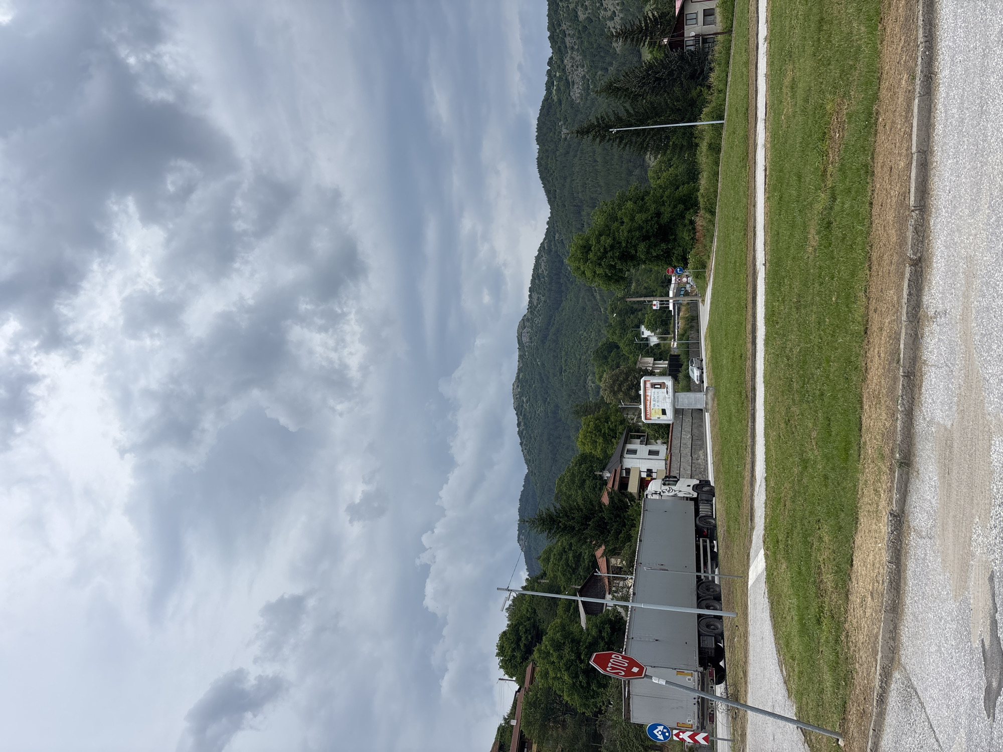 This is a vertical, eye-level shot of a rural Bulgarian scene under a cloudy sky. The foreground shows a paved road with a white line marking its edge, followed by a strip of green grass and then a curb. A large, gray semi-trailer truck is parked on the side of the road, facing away from the camera. A red octagonal "STOP" sign is visible on a pole near the truck, along with a traffic sign indicating a sharp curve to the right.
Behind the truck, a small cluster of houses and trees are nestled at the base of a steep, tree-covered mountain. The mountain dominates the background, its slopes shrouded in a dense green forest. The sky is overcast with heavy, gray clouds, suggesting an impending storm or recent rain. The overall atmosphere is somewhat somber due to the weather. A billboard with some text is visible near the top of the frame, on the right side, and a person appears to be near a small white building in the distance. The scene is static, with no apparent activity other than the parked truck. The time of day is unclear due to the overcast conditions, but it appears to be daytime.