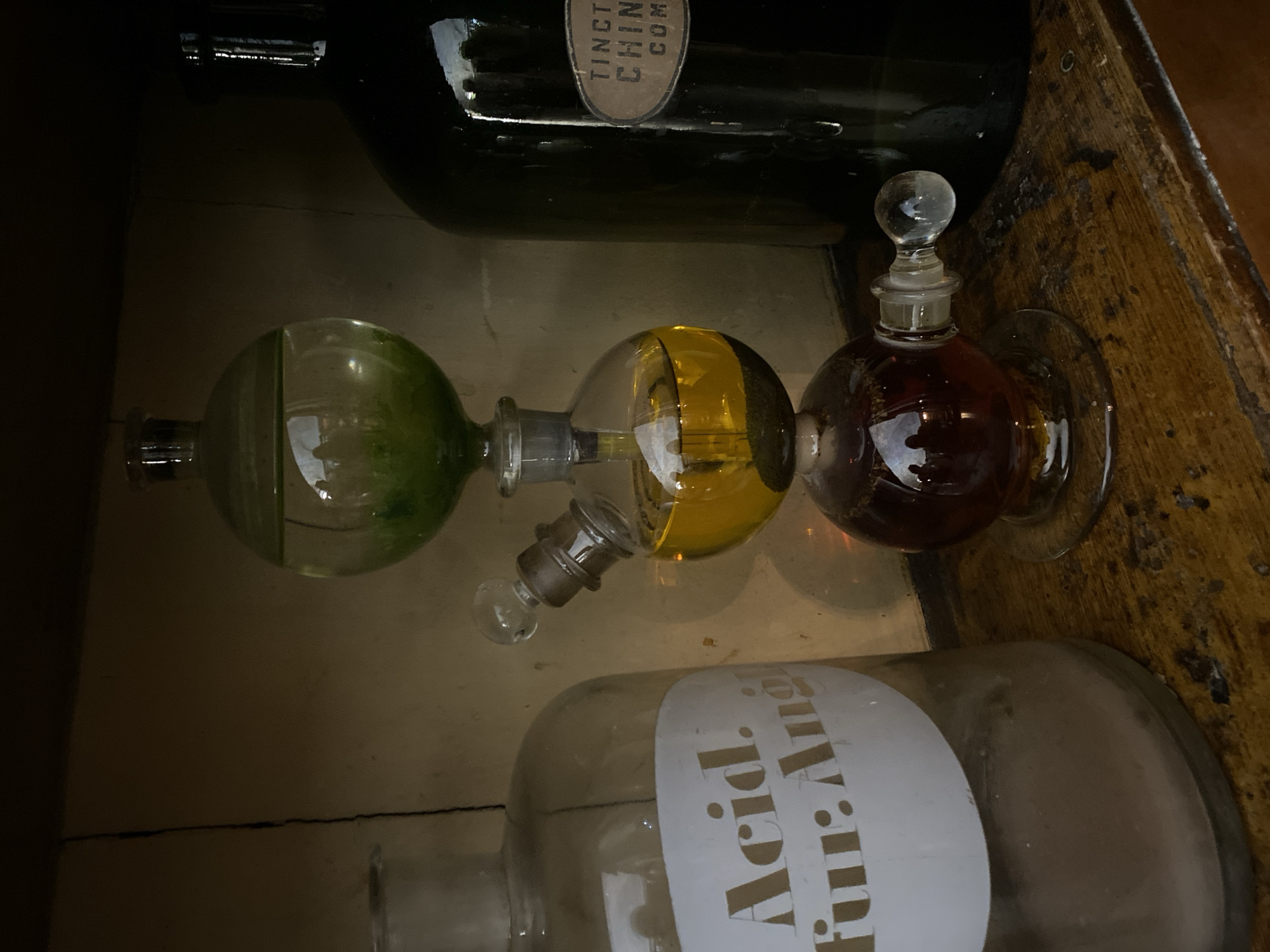 The image depicts a still life of old apothecary or laboratory equipment displayed on a wooden shelf. In the center is a three-tiered glass apparatus containing liquids of different colors: green in the top bulb, yellow in the middle bulb, and dark amber in the bottom bulb. The apparatus has stoppers and connections made of glass. To the left is a large, clear glass bottle with a label that reads "Acid. fur: Aug.". To the right is a dark green glass bottle with a label that reads "TINCT. CHIN CON". The overall impression is one of historical scientific or medicinal items, possibly in a museum or an antique shop. The background appears to be a wooden shelf or cabinet. There are reflections of people in the glass bottles, suggesting the photograph was taken in an indoor setting with lighting.
