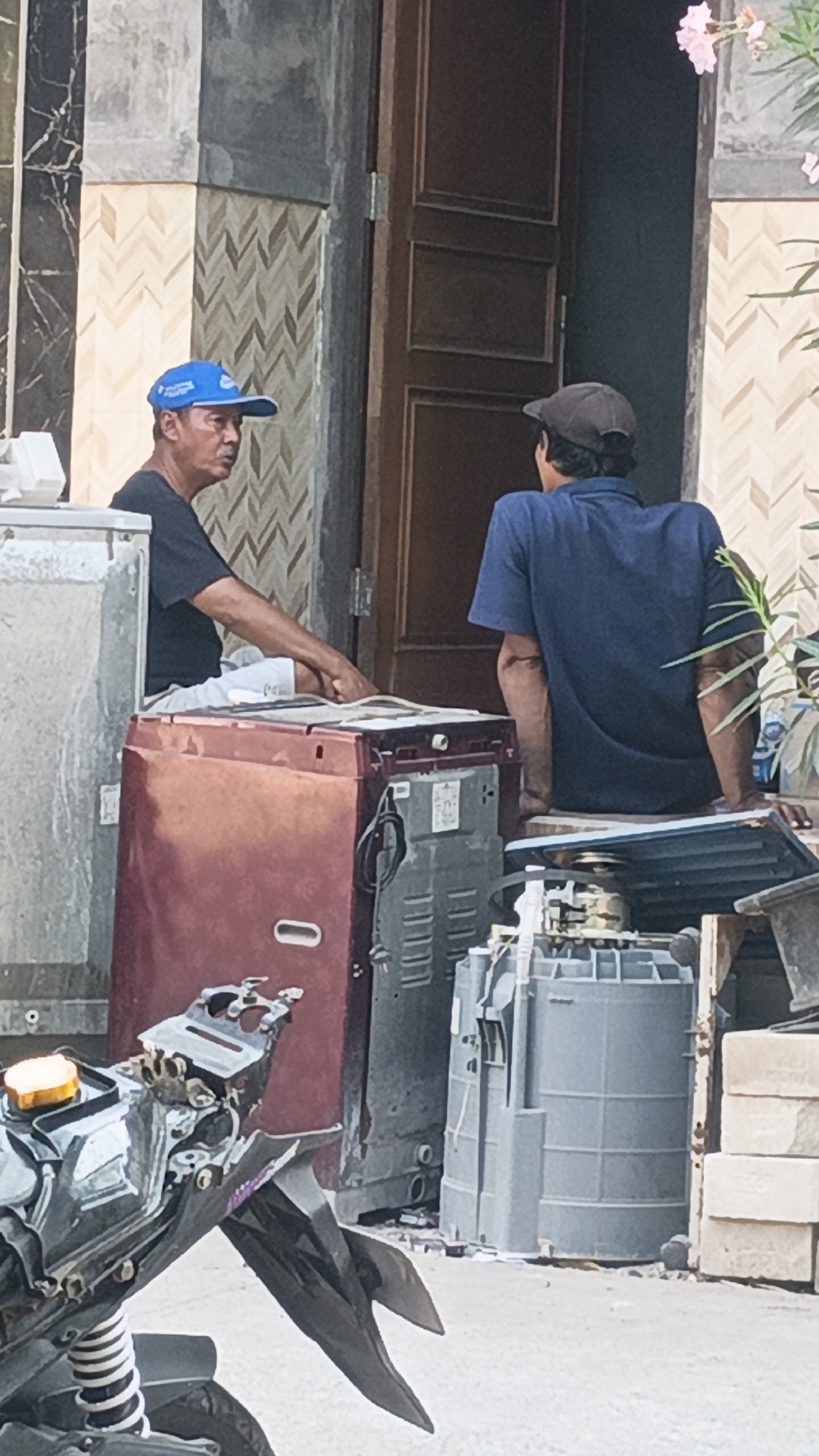 Two men, one wearing a blue cap and the other a dark cap, are sitting in front of a doorway. The man with the blue cap is seated on a maroon washing machine and is looking to his left. The man with the dark cap is seated on a piece of wood in front of a large gray cylindrical object, facing away from the camera. In the foreground, a motorcycle is partially visible. The background features a building with a tiled facade and a wooden door. The scene appears to be located outdoors, possibly in a residential or workshop area, in Bulungcangkring, Indonesia.