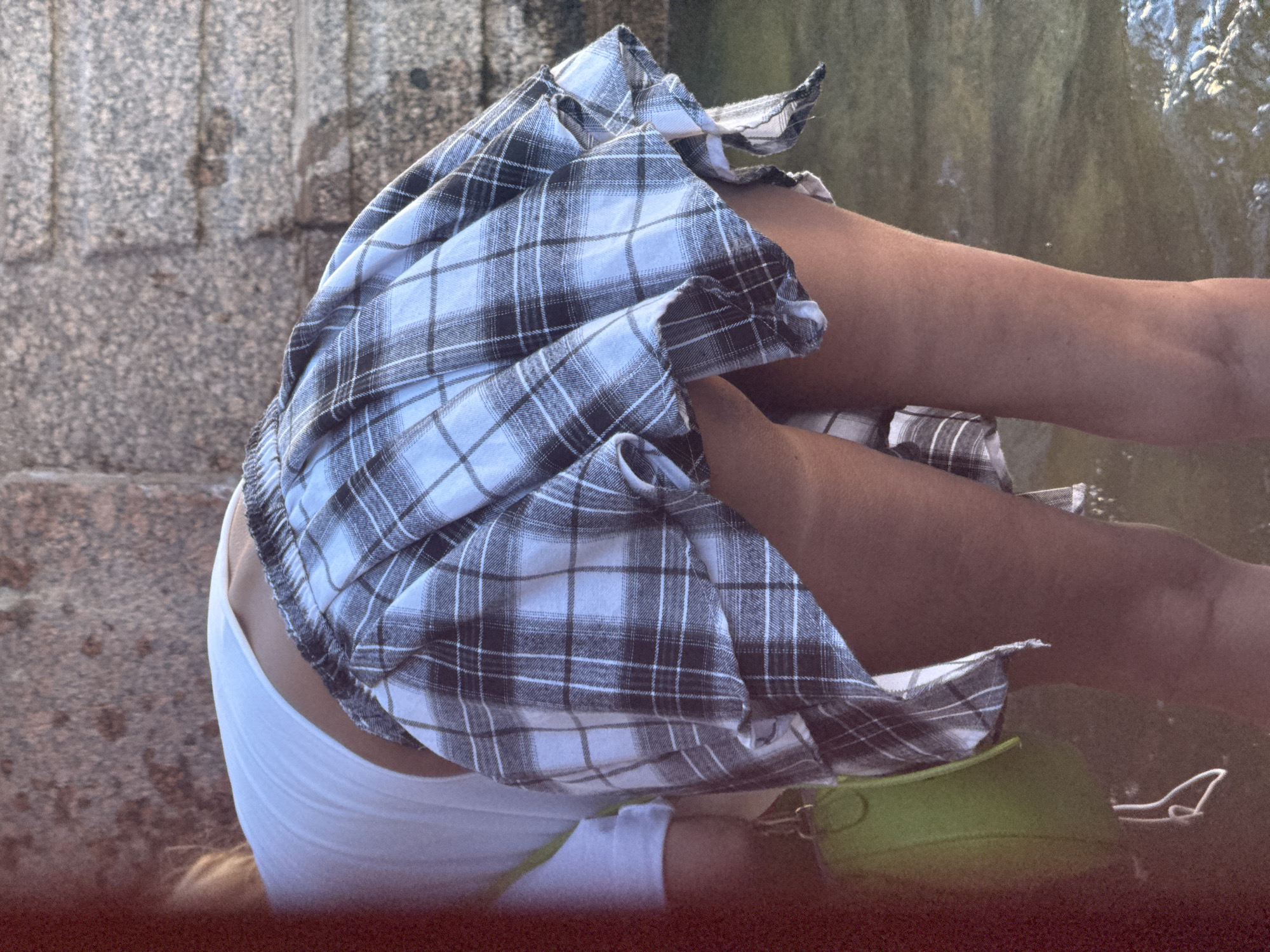 This is a medium-shot, low-angle photograph of a person sitting with their legs exposed and their skirt lifted. The subject is wearing a white tank top and a blue and white plaid skirt. Their legs are tanned and their knees are bent. A bright green purse with a strap is visible next to their legs. In the background, a textured gray stone wall is on the left, and a body of water with reflections of light is on the right. The lighting suggests it is daytime, possibly late afternoon or early evening, with the sun casting warm tones. The scene appears to be outdoors, near a fountain or body of water, perhaps in a park or public space within Saint Petersburg, Russia, given the stone architectural style. The overall mood is casual and candid, capturing a fleeting moment. No text is visible in the image.